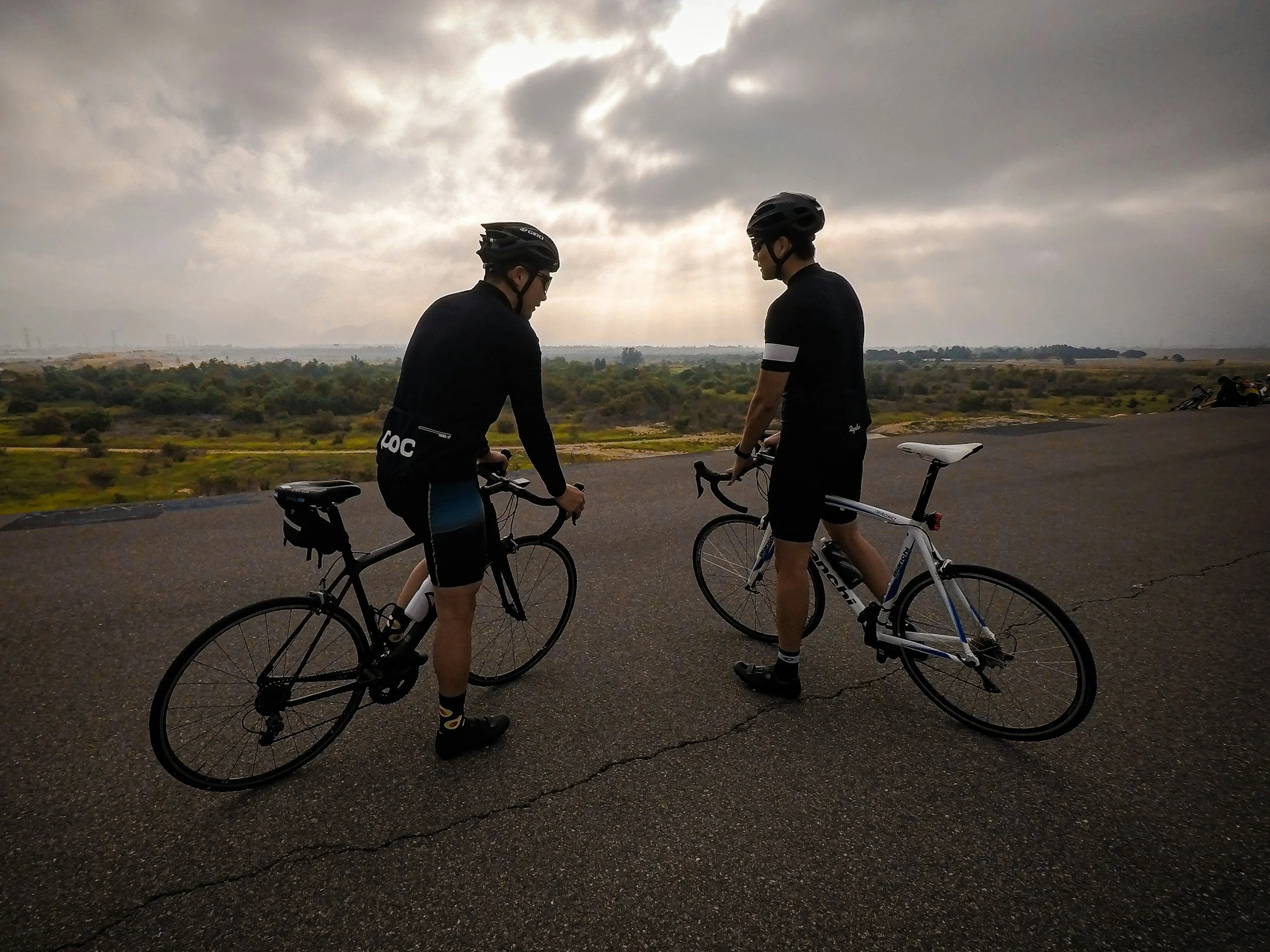 Two friendly cyclists wearing with their road bikes , black helmets and cycling gear stand on a paved road, facing each other, with a scenic landscape and cloudy sky in the background.