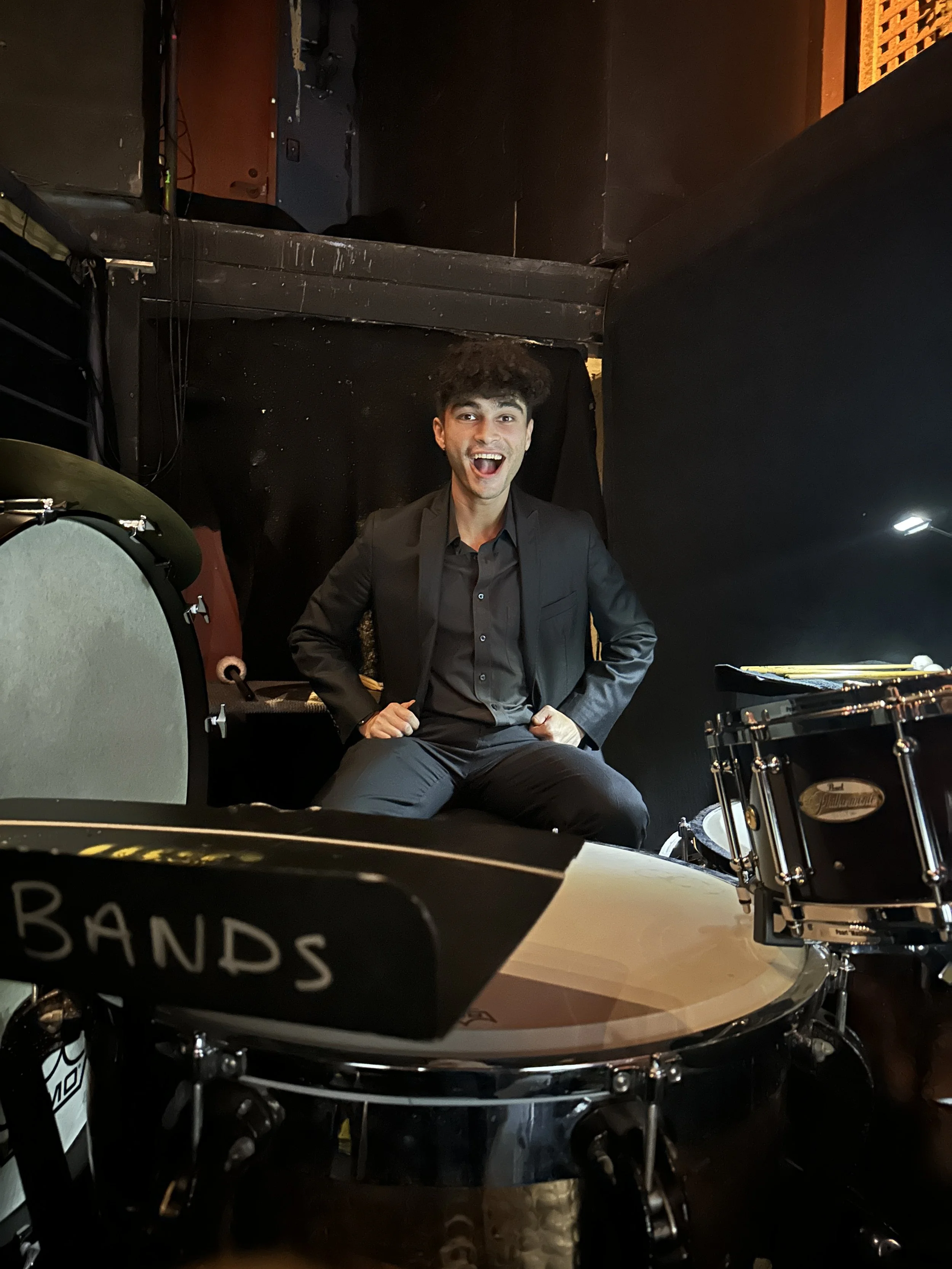 A man in a black suit sitting on a drum set with a big smile, in a dark indoor setting with drums and musical equipment around him.
