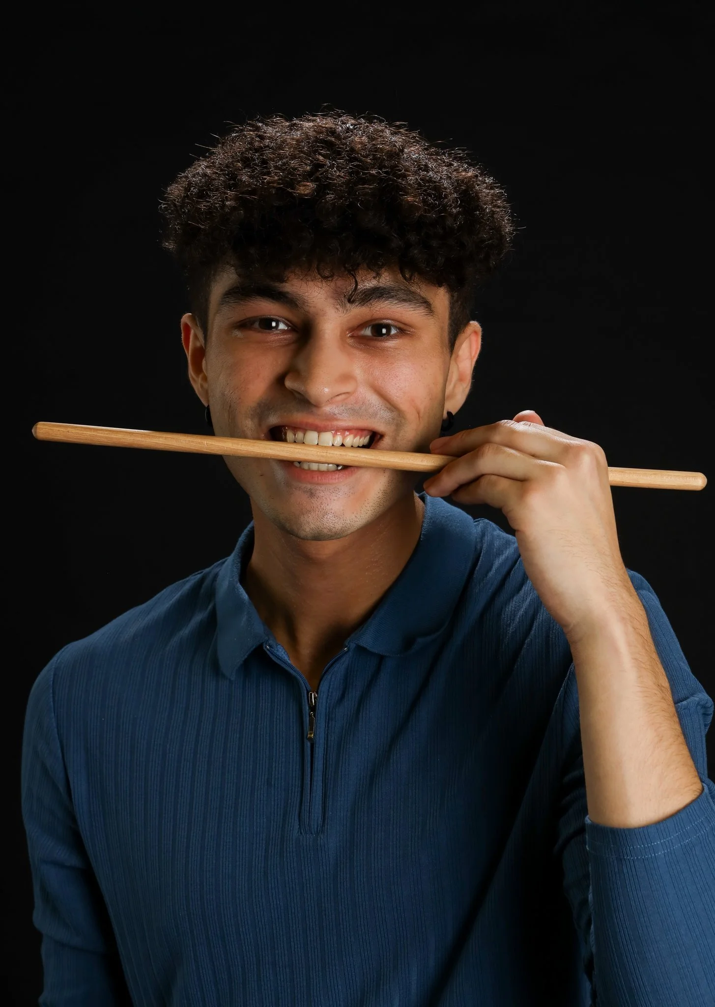 A smiling man with curly hair holding a chopstick in his mouth against a black background.