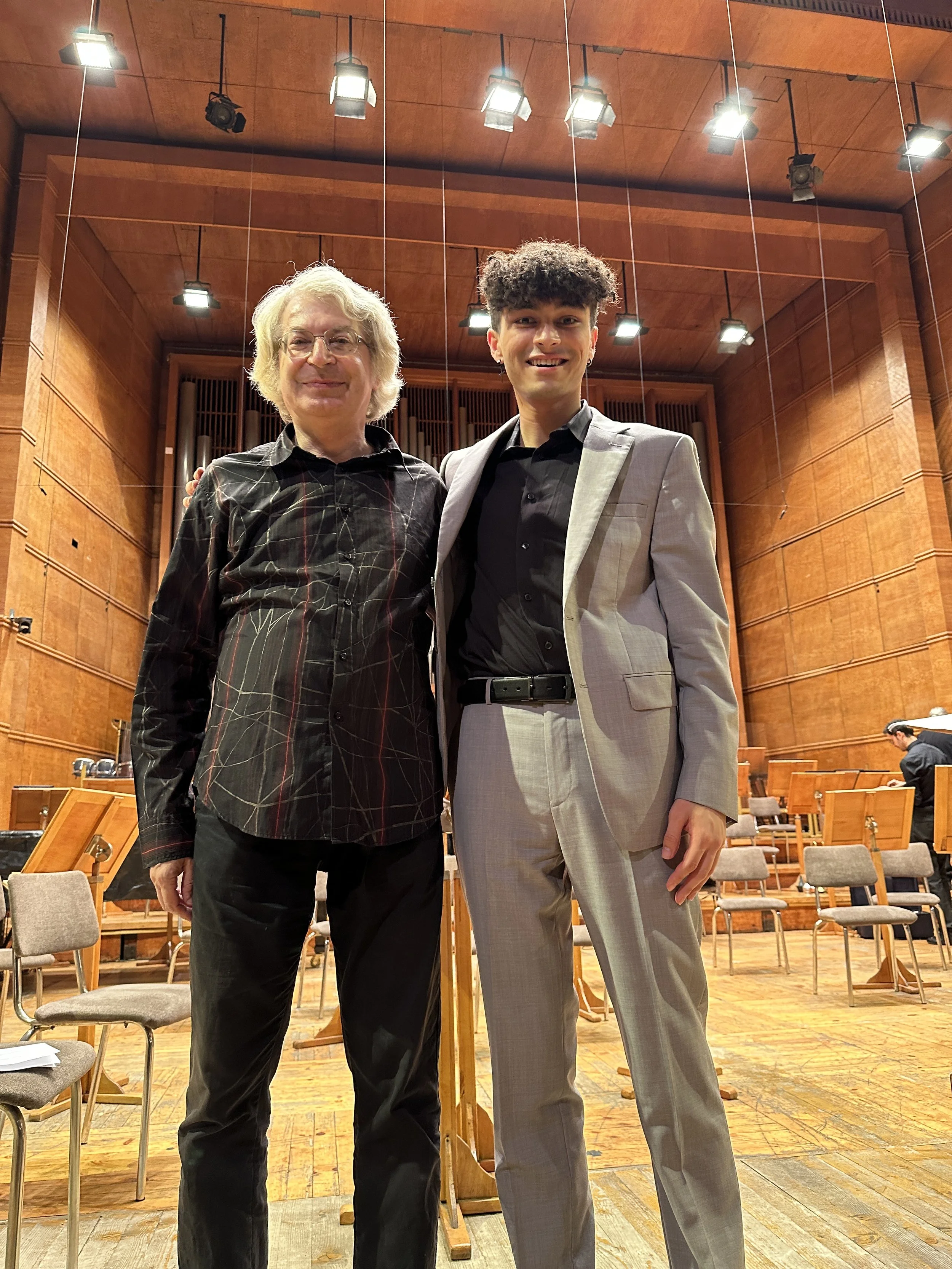 Two men standing together on stage in a concert hall, smiling at the camera. The man on the left has gray hair, glasses, and is wearing a black shirt with colorful lines, while the man on the right has curly dark hair and is wearing a light gray suit