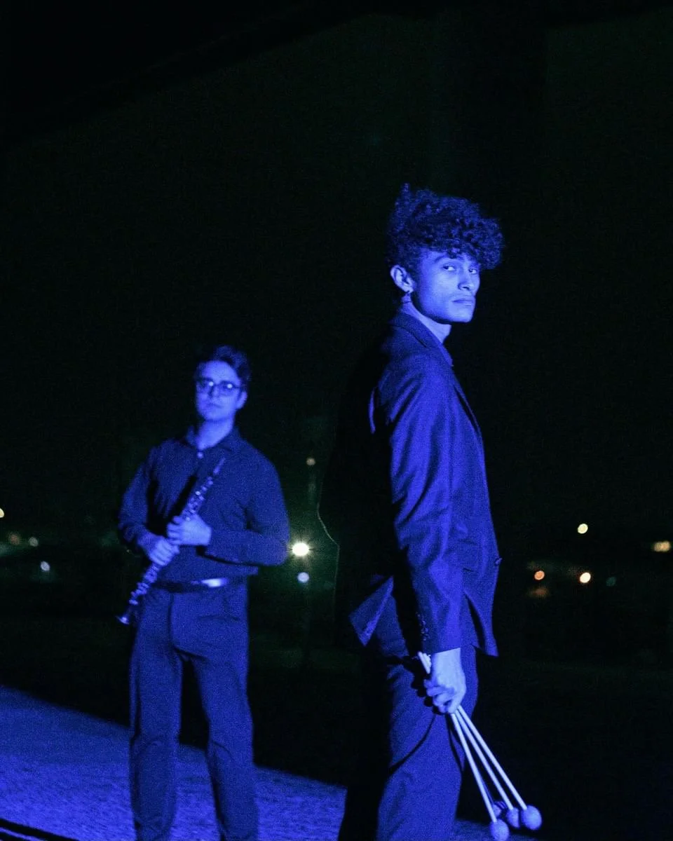 A young man and woman stand outside at night under a dark sky, illuminated by blue lighting. The woman is holding a clarinet and the man is holding a pair of tambourines. They are dressed in dark clothing and look serious.