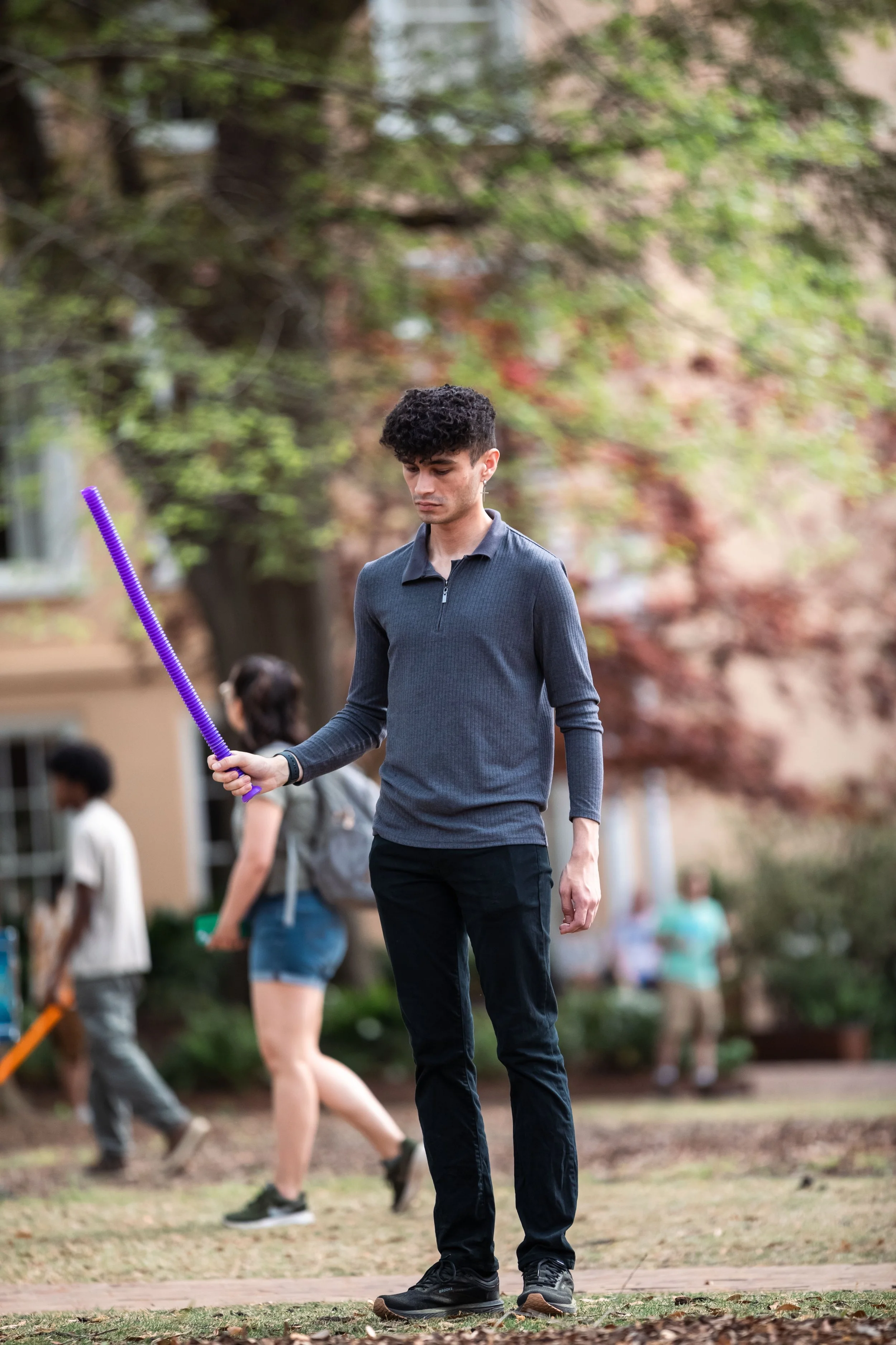 A young man standing outdoors holding a purple stick, with other people in the background in a park setting with trees and buildings.