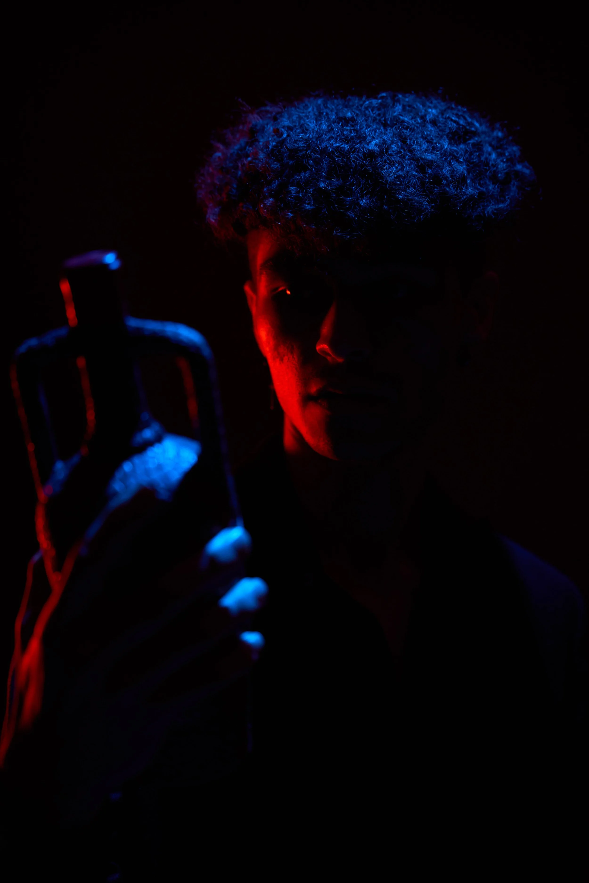 A person with curly hair holding a bottle, illuminated with red and blue lighting in a dark setting.