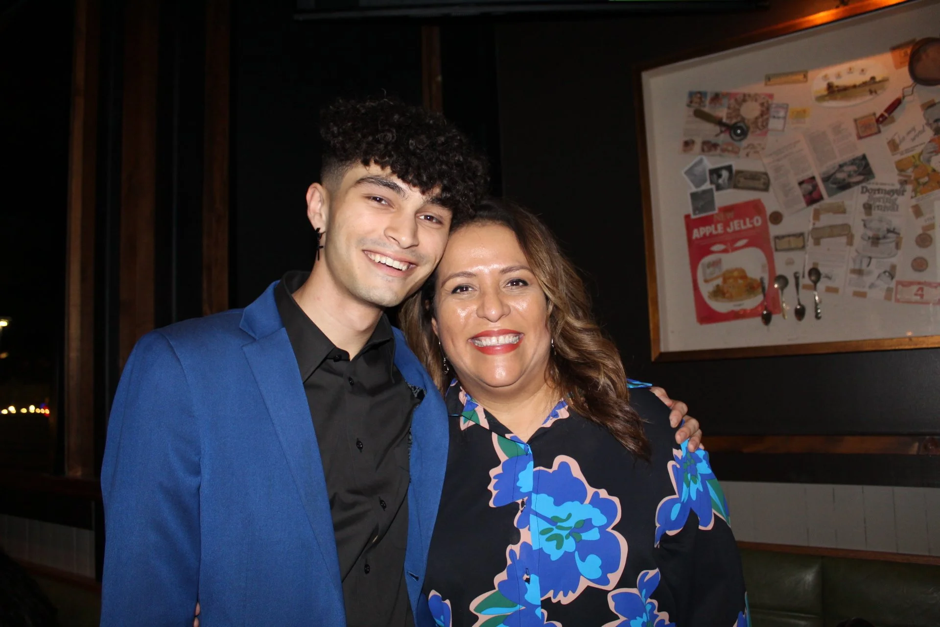 A young man and an older woman smiling and posing together in a dimly lit indoor setting, with a bulletin board in the background.