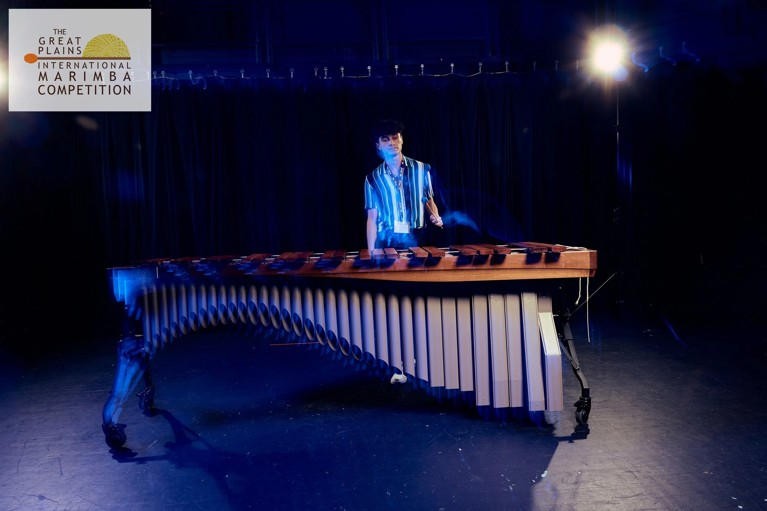 A person standing behind a marimba on a stage with dark curtains in the background and stage lighting overhead.