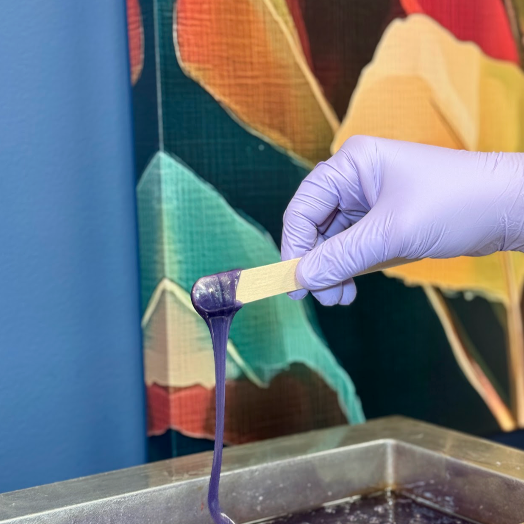 A gloved hand dipping a stick into purple slime in a tray, with colorful geometric artwork in the background.