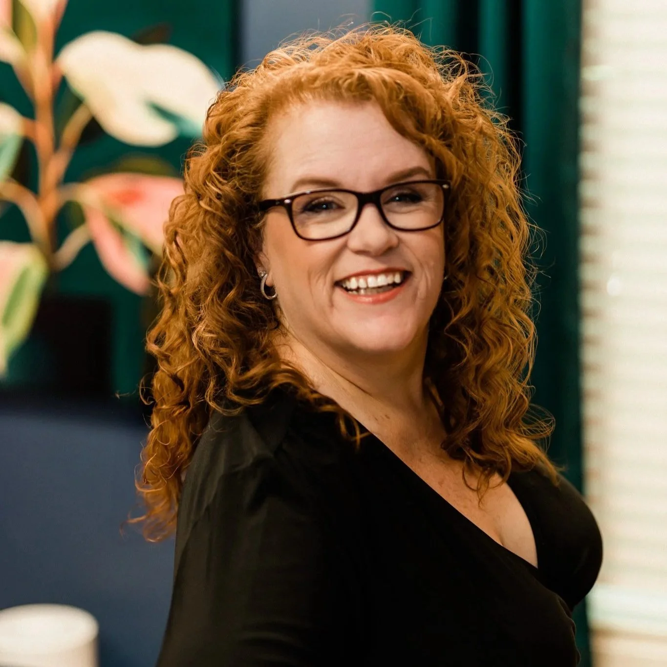 A smiling woman with curly red hair, wearing glasses and a black top, standing indoors with plants and window blinds in the background.