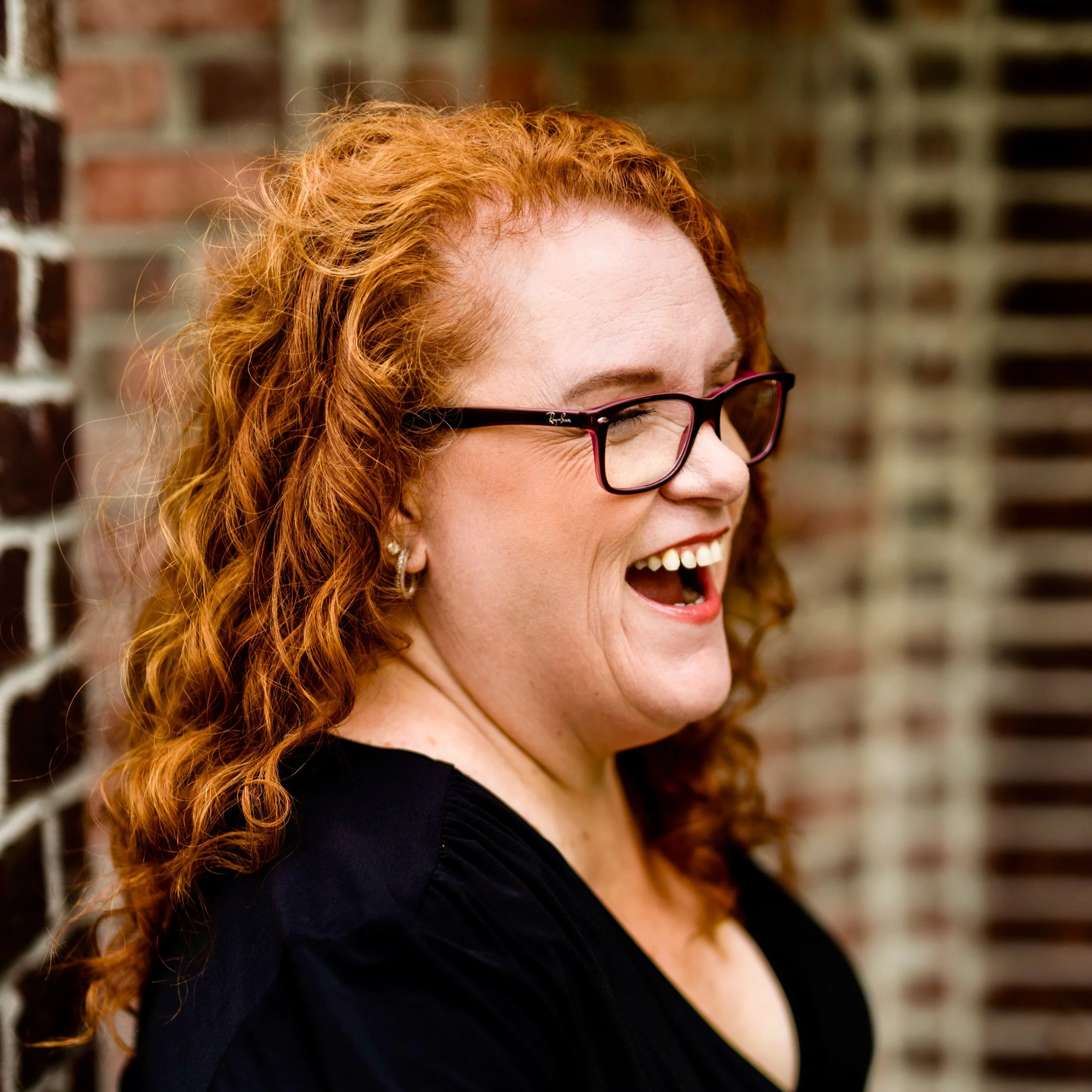 A woman with curly red hair and glasses laughing outdoors against a brick wall.