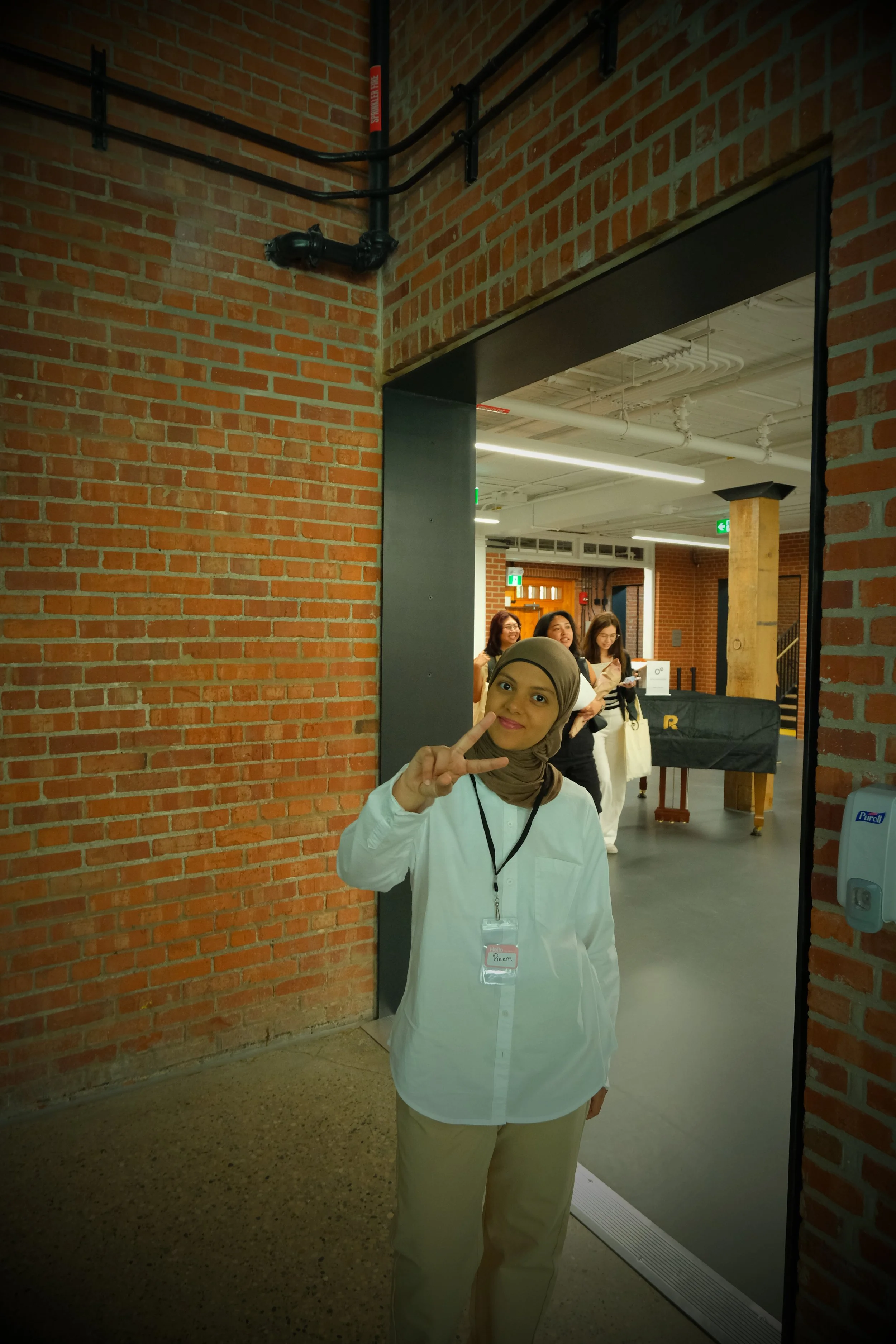 Woman wearing a hijab and white shirt making a peace sign in front of a mirror, with a group of women in the background inside a modern industrial-style space with brick walls and exposed pipes.