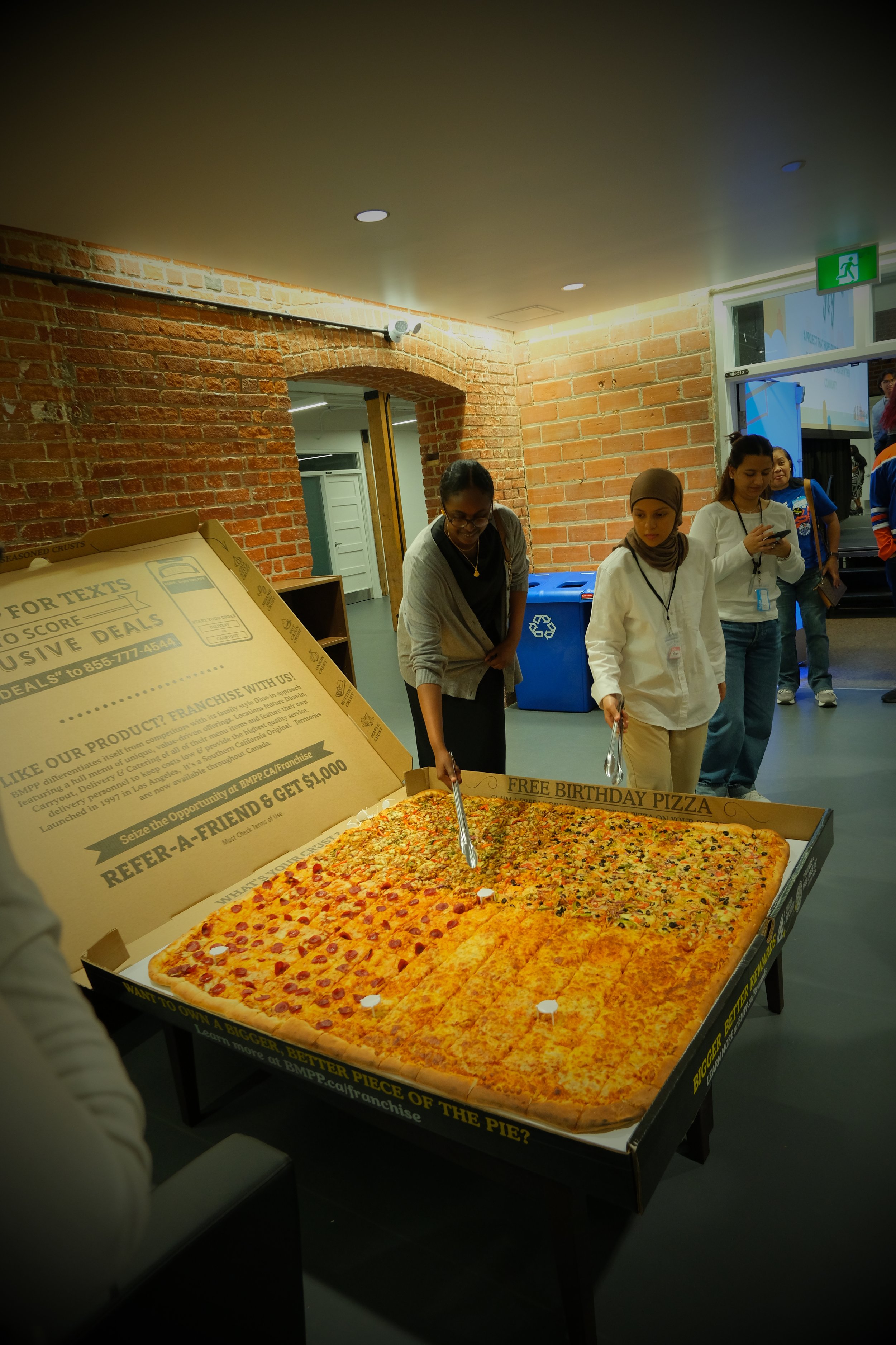 People gathered around a large pizza in a cardboard box, inside a brick-walled room.