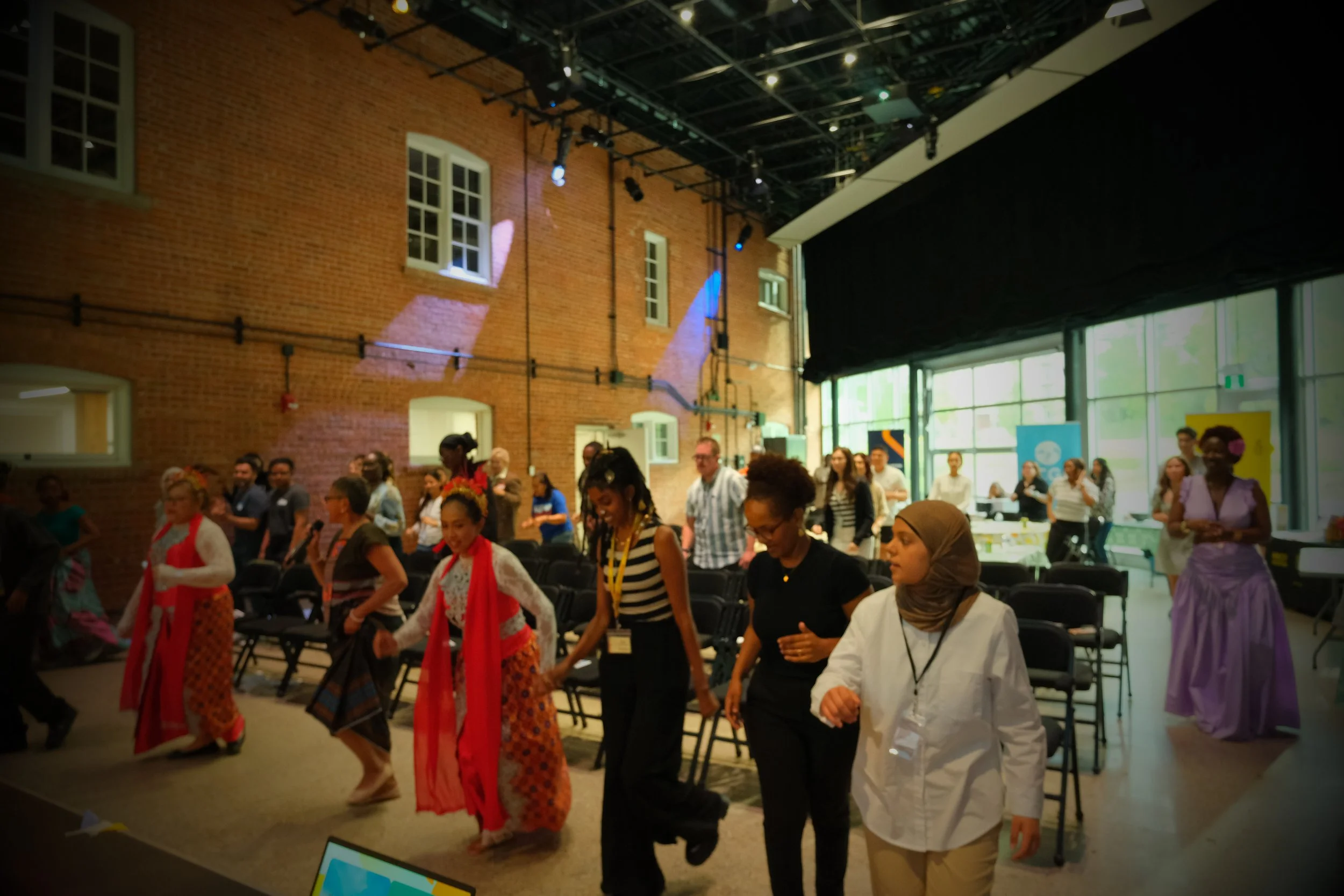 People participating in a cultural dance event indoors, dressed in traditional and contemporary clothing, with a brick wall and large windows in the background.