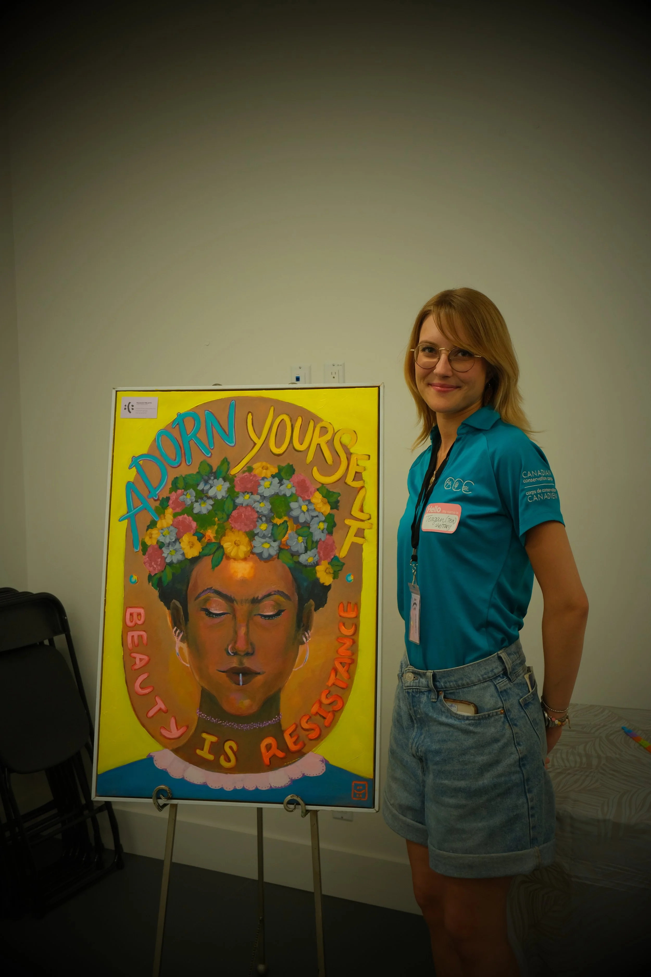 A smiling woman with glasses and blonde hair standing next to a colorful illustration of a woman with a flower crown and the text 'ADORN YOURSELF, BEAUTY IS RESISTANCE' in a bright room.