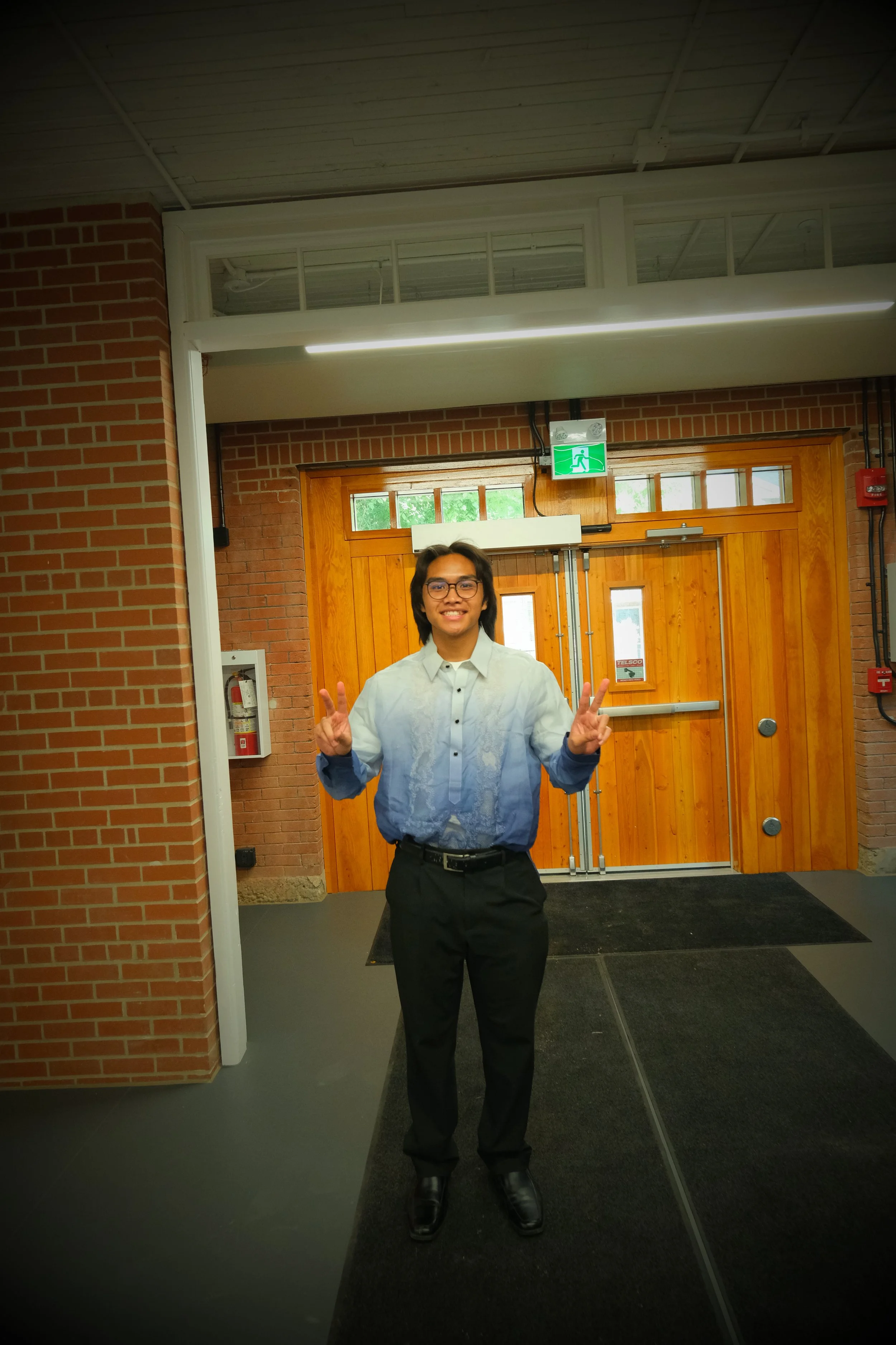 Young man standing inside near a wooden door, smiling and making peace signs with both hands.