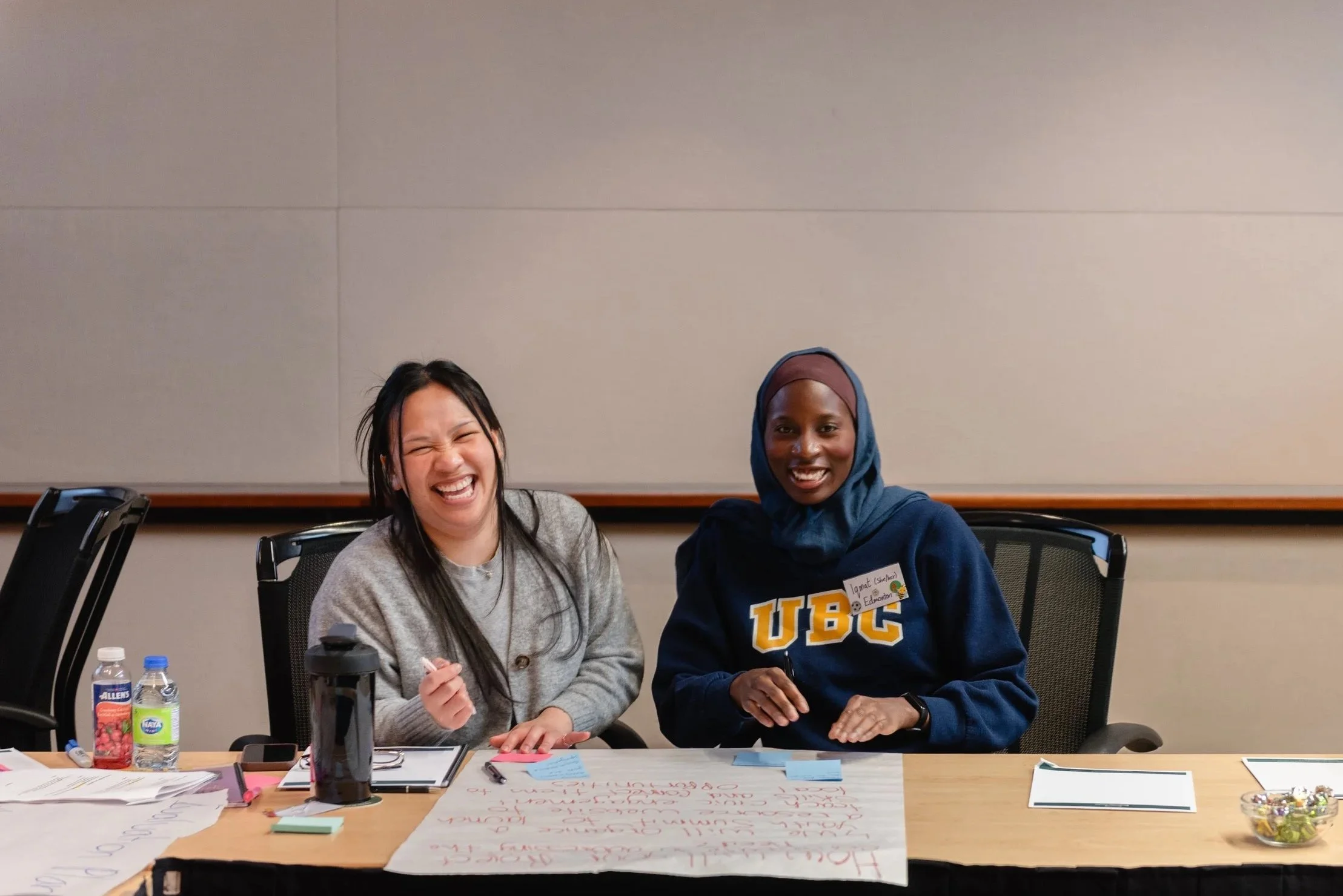 Two women sitting at a conference table, laughing. One is wearing a gray sweater, the other a blue hoodie with 'UBC' printed. The table has papers, sticky notes, a water bottle, and a bowl of candies.