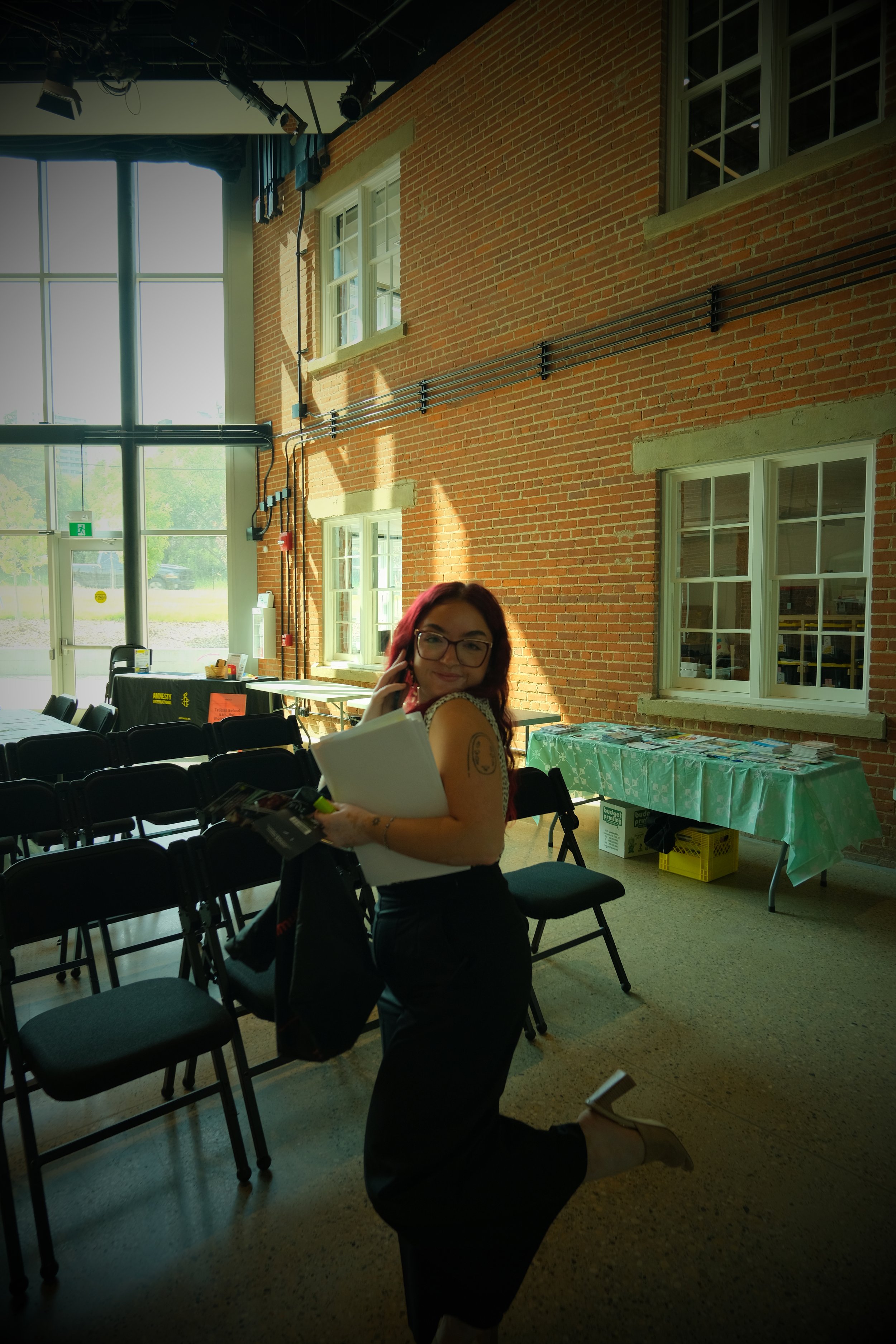 A young woman with reddish hair and glasses smiling while holding papers, standing in a sunlit room with large windows and a brick wall, surrounded by chairs and tables.