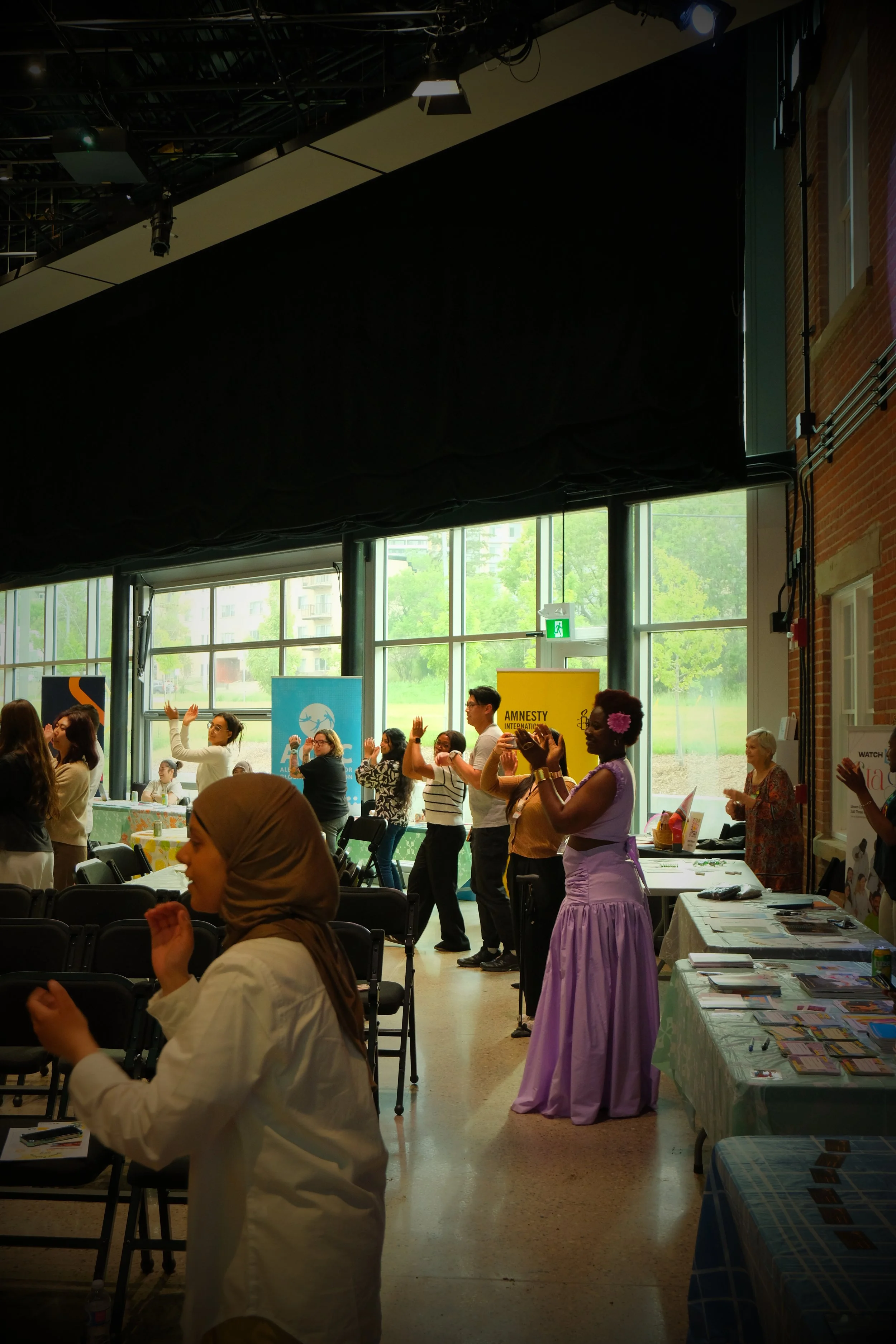 People participating in a dance or exercise workshop indoors, with banners and tables displaying informational material, near large windows with greenery outside.