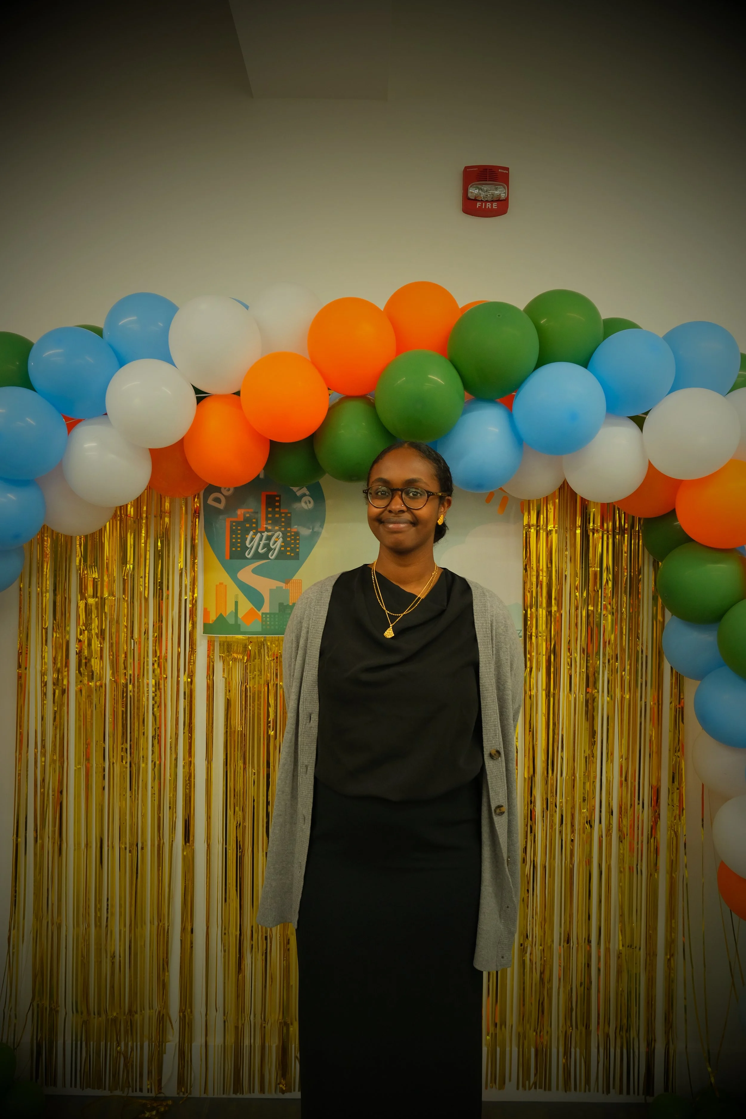 A woman in a black dress and gray cardigan standing in front of a colorful balloon arch with gold fringe curtains and a cityscape banner, celebrating Diversity and Equity.