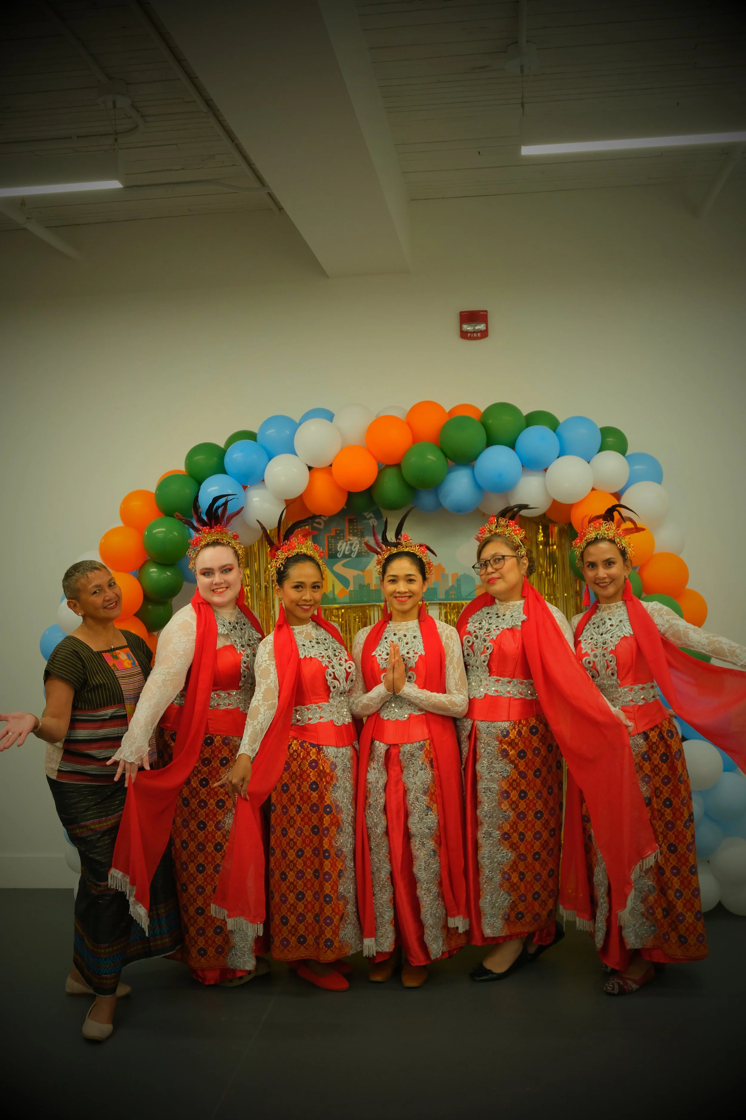 Group of six women in traditional Southeast Asian attire standing in front of a balloon arch with orange, white, blue, and green balloons, posing for a photo at a cultural event.