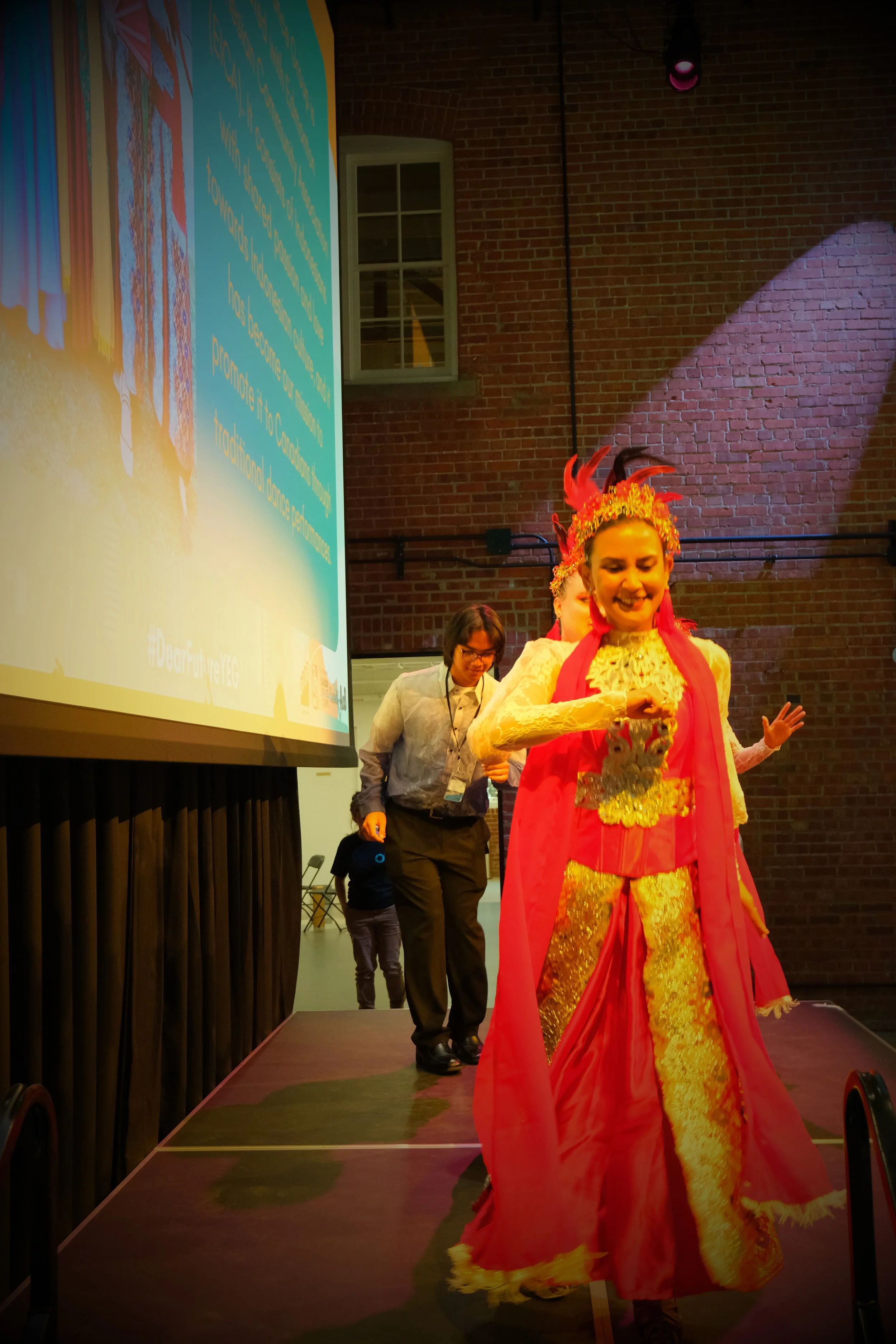 Women dressed in traditional Asian costume with pink and gold colors, walking on stage with a brick wall background and a large projector screen displaying text.