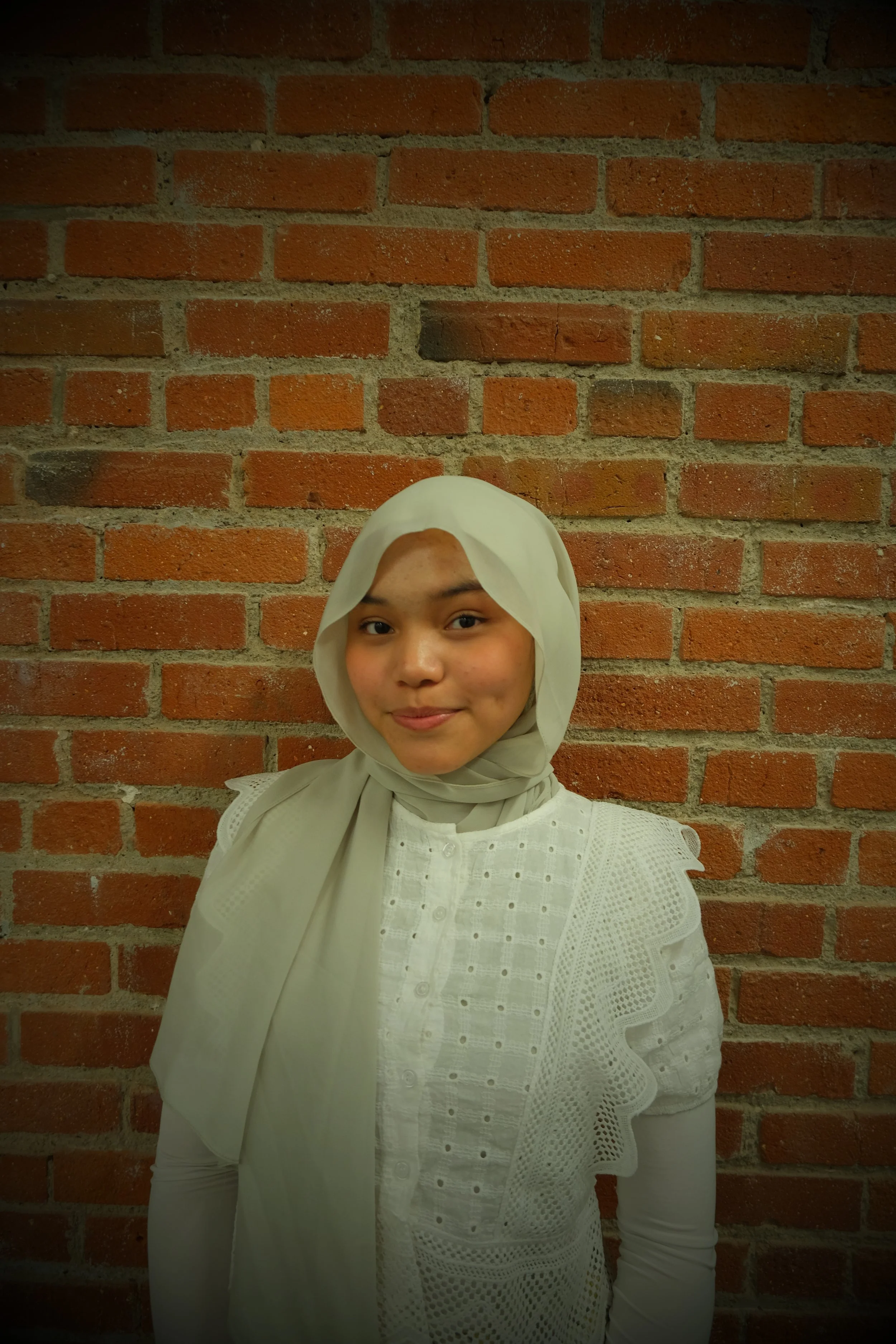 A young woman with a beige hijab and white embroidered blouse standing in front of a brick wall.