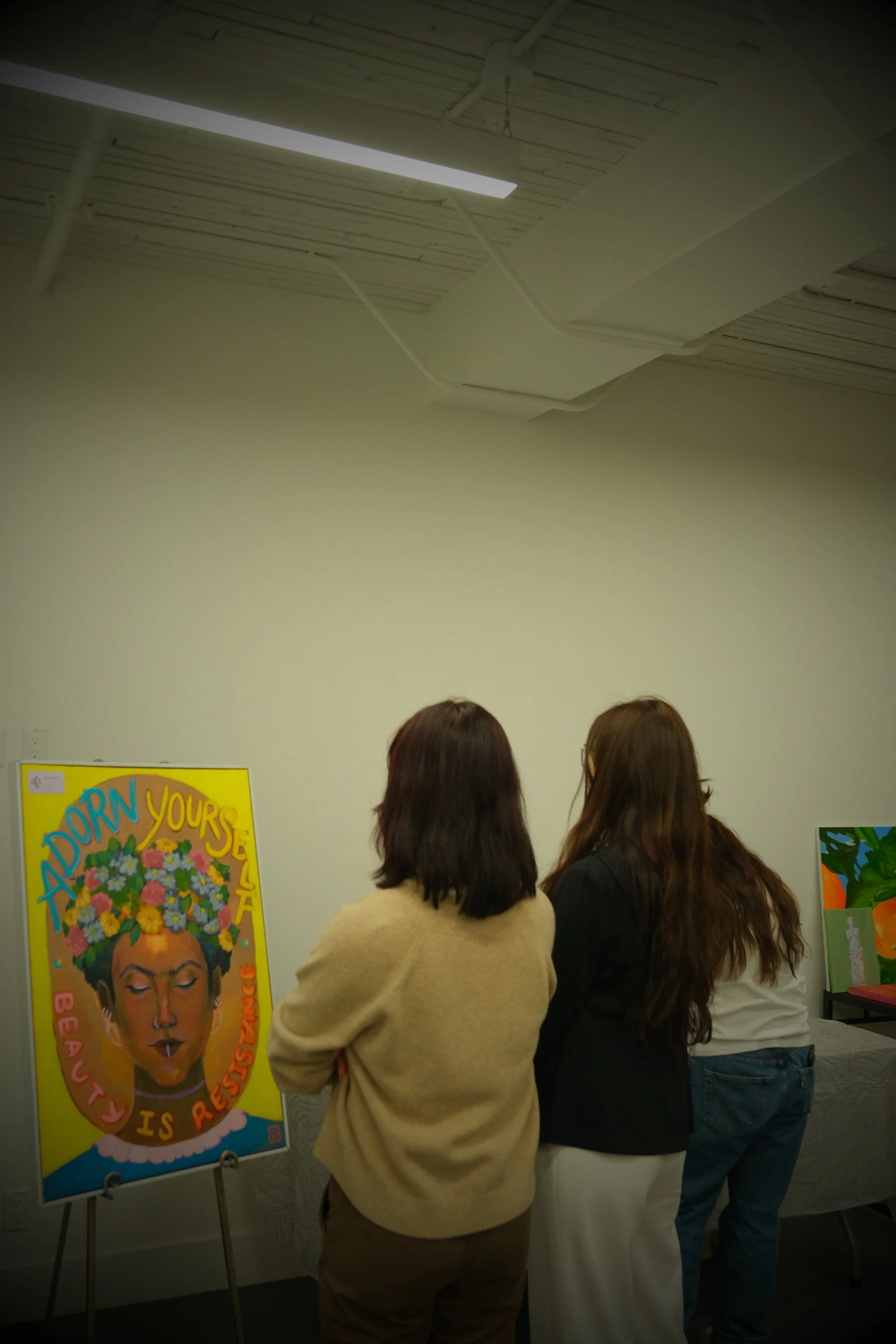 Three women observing a colorful portrait of a woman with flowers in her hair, and the message "A DORN YOURSELF BE A BEAUTY IS RESISTANCE," displayed on an easel in an art gallery or exhibition space.