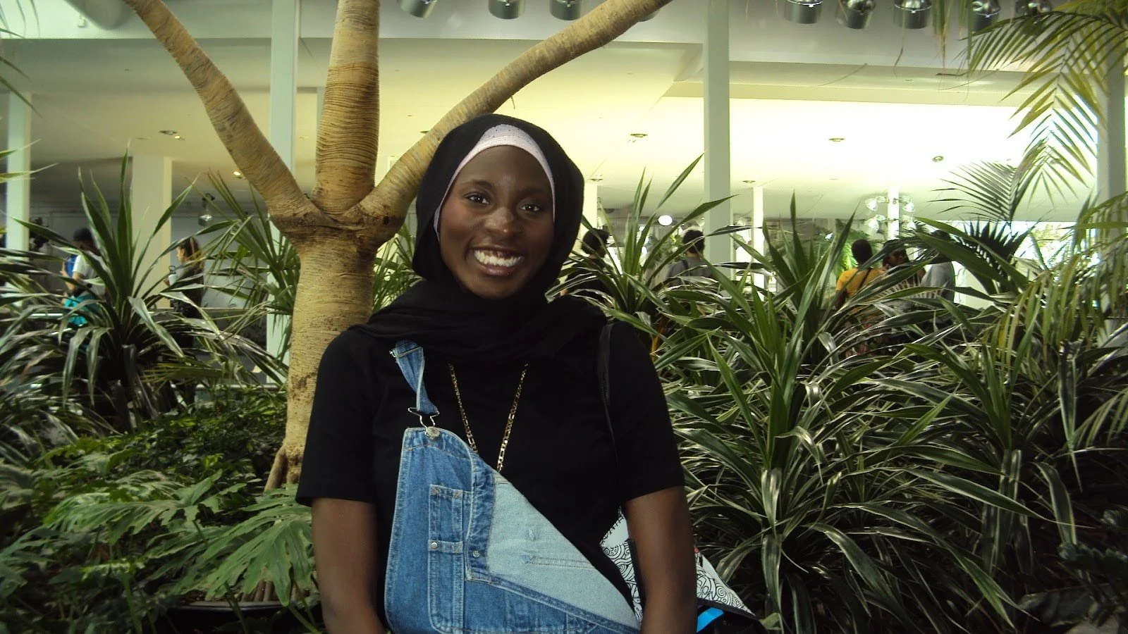 Smiling woman with dark skin and a headscarf, standing in front of a tree and indoor plants, in a modern well-lit indoor setting.