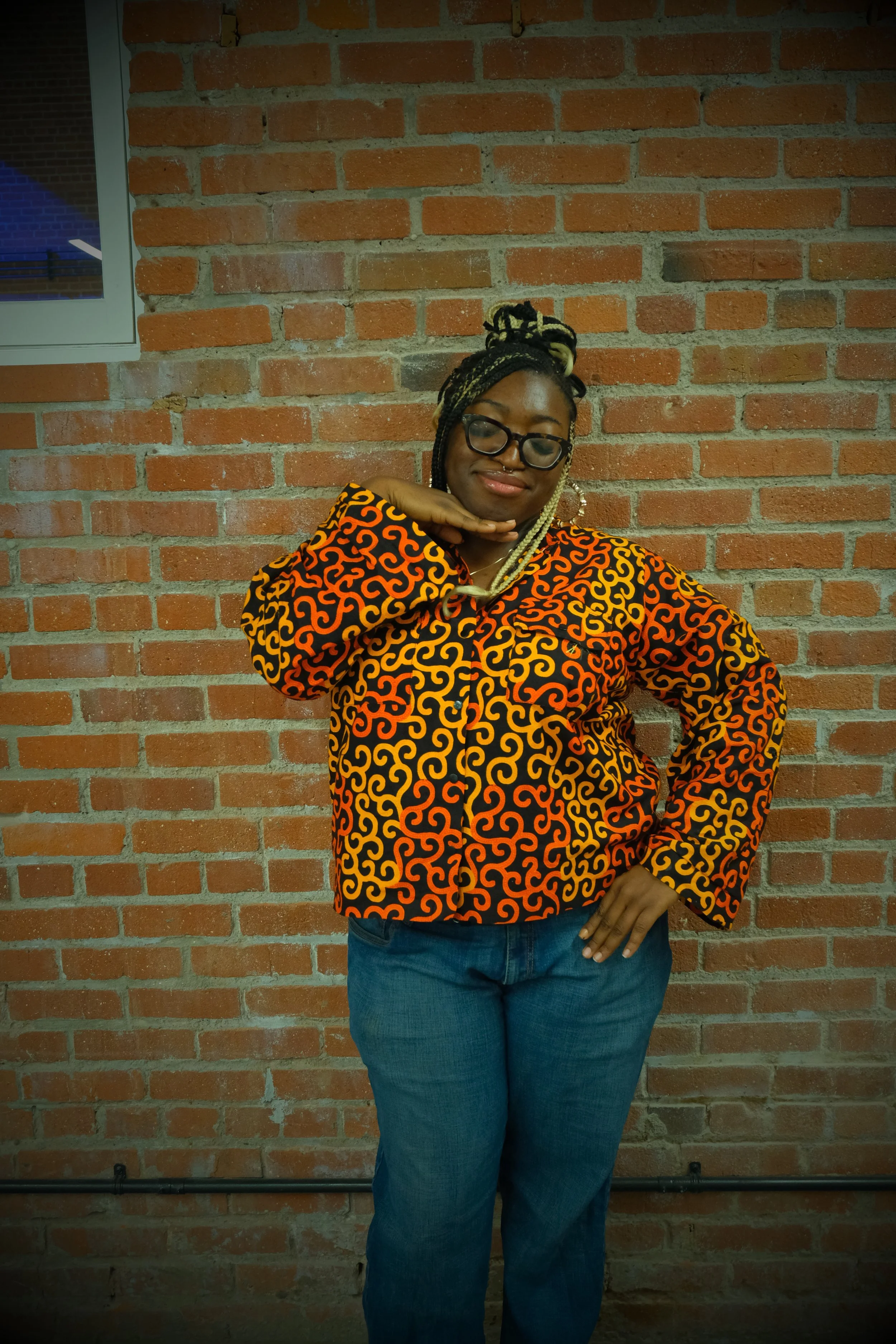 A woman with braided hair and glasses is standing in front of a brick wall, wearing a black and orange patterned jacket, blue jeans, and striking a pose with one hand on her hip and the other hand under her chin.