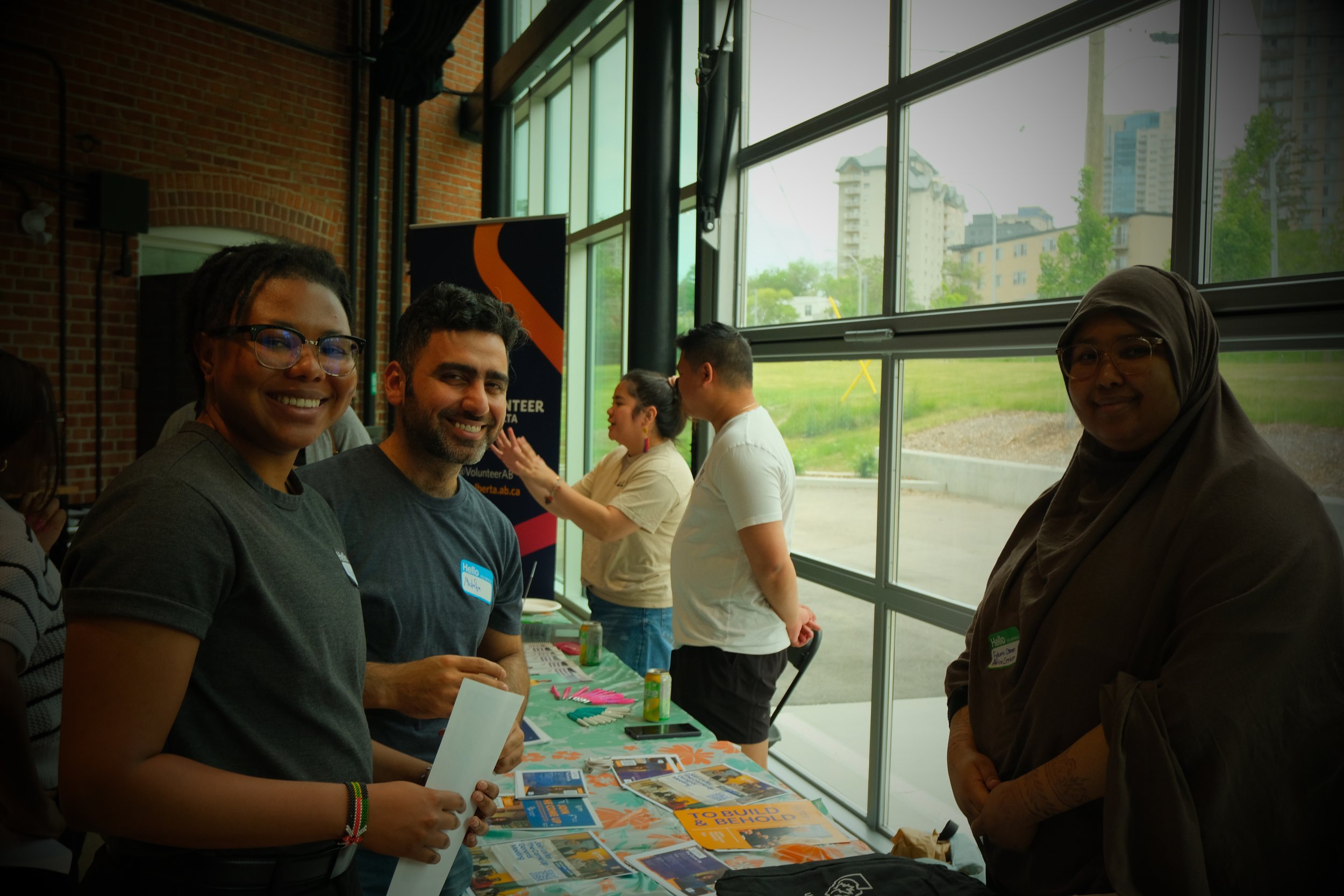 People standing behind a table at an indoor event, engaging with each other, with brochures and canned drinks on the table, and large windows showing buildings and greenery outside.