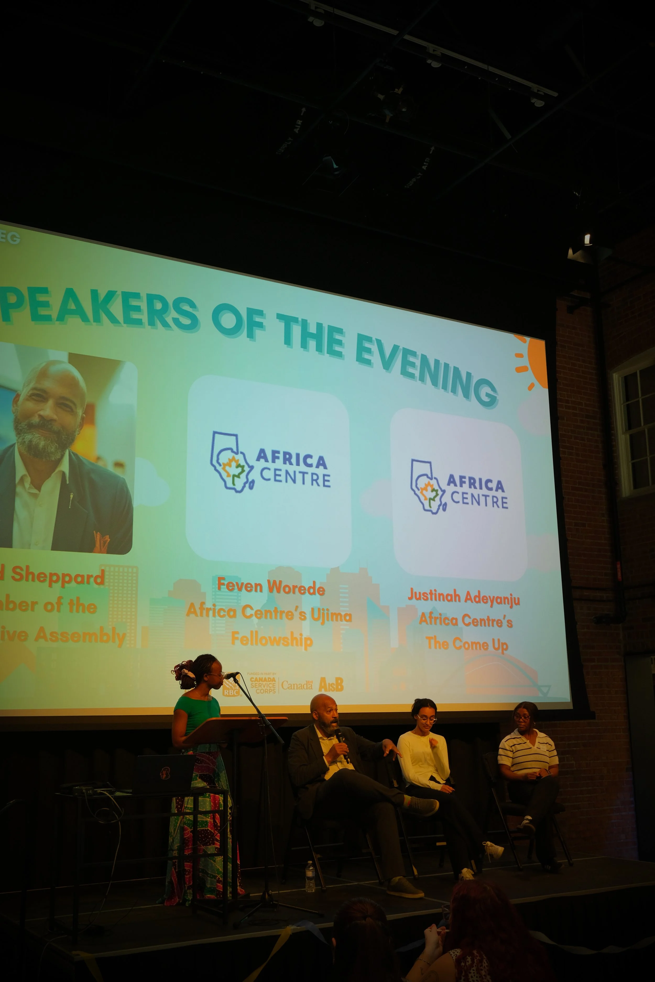 A panel discussion at an event with people sitting on stage in front of a large screen displaying information about speakers and organizations involved, including Africa Centre. Four people are on stage, one standing at a podium and three seated with microphones.