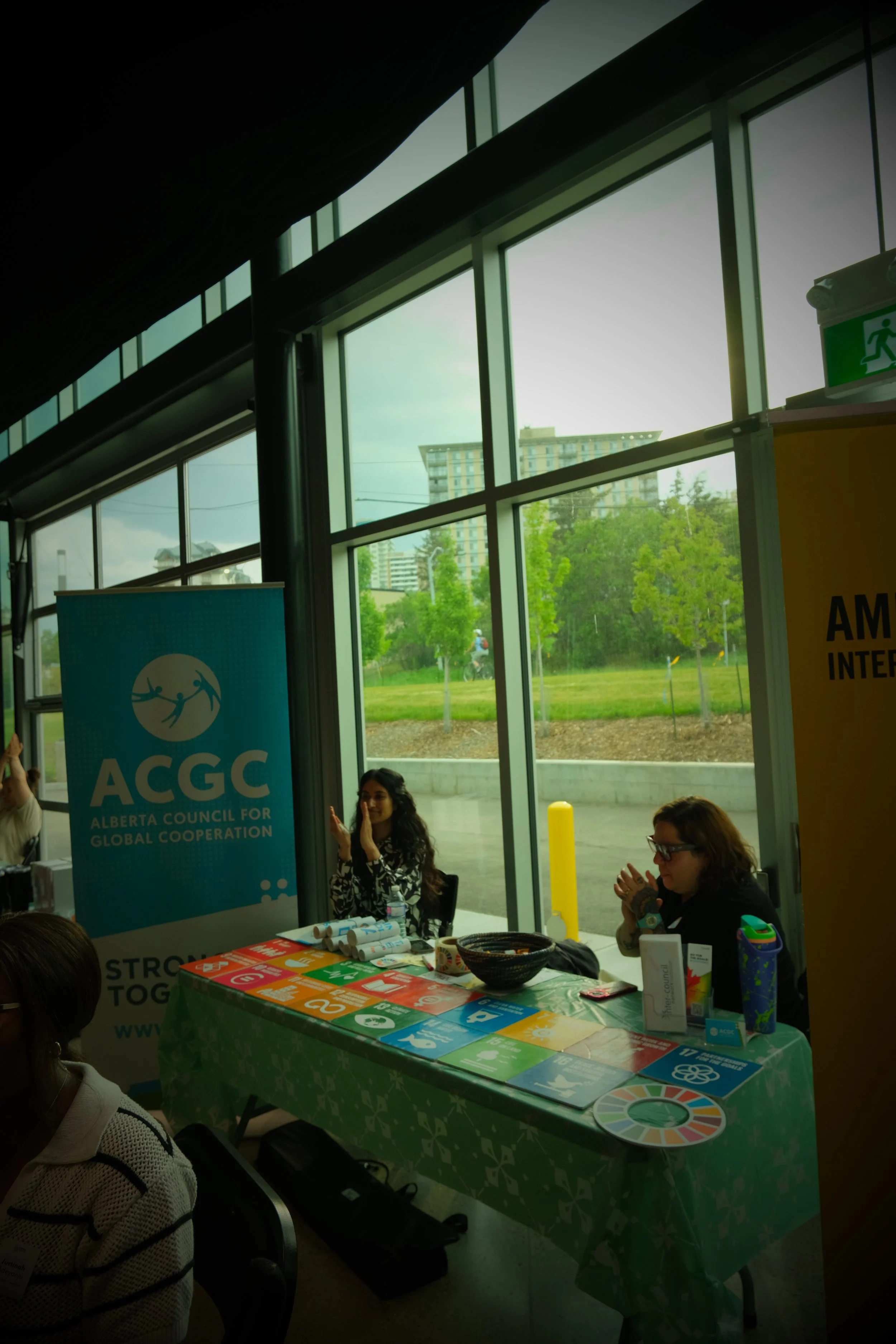 Indoor booth with two women sitting at a table covered with colorful cards and materials, near large windows showing trees and buildings outside, with a banner reading 'ACGC Alberta Council for Global Cooperation'.