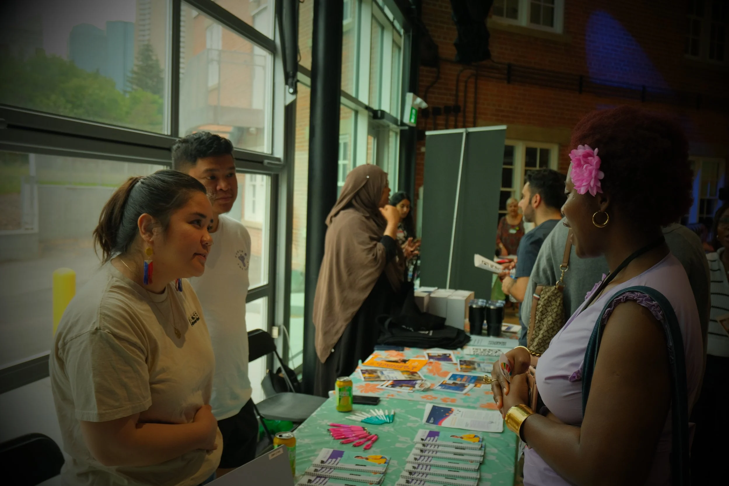 People at an indoor event booth, engaging in conversations. A woman with a pink flower in her hair is talking to two young people behind the booth, which displays pamphlets, flyers, and promotional items.