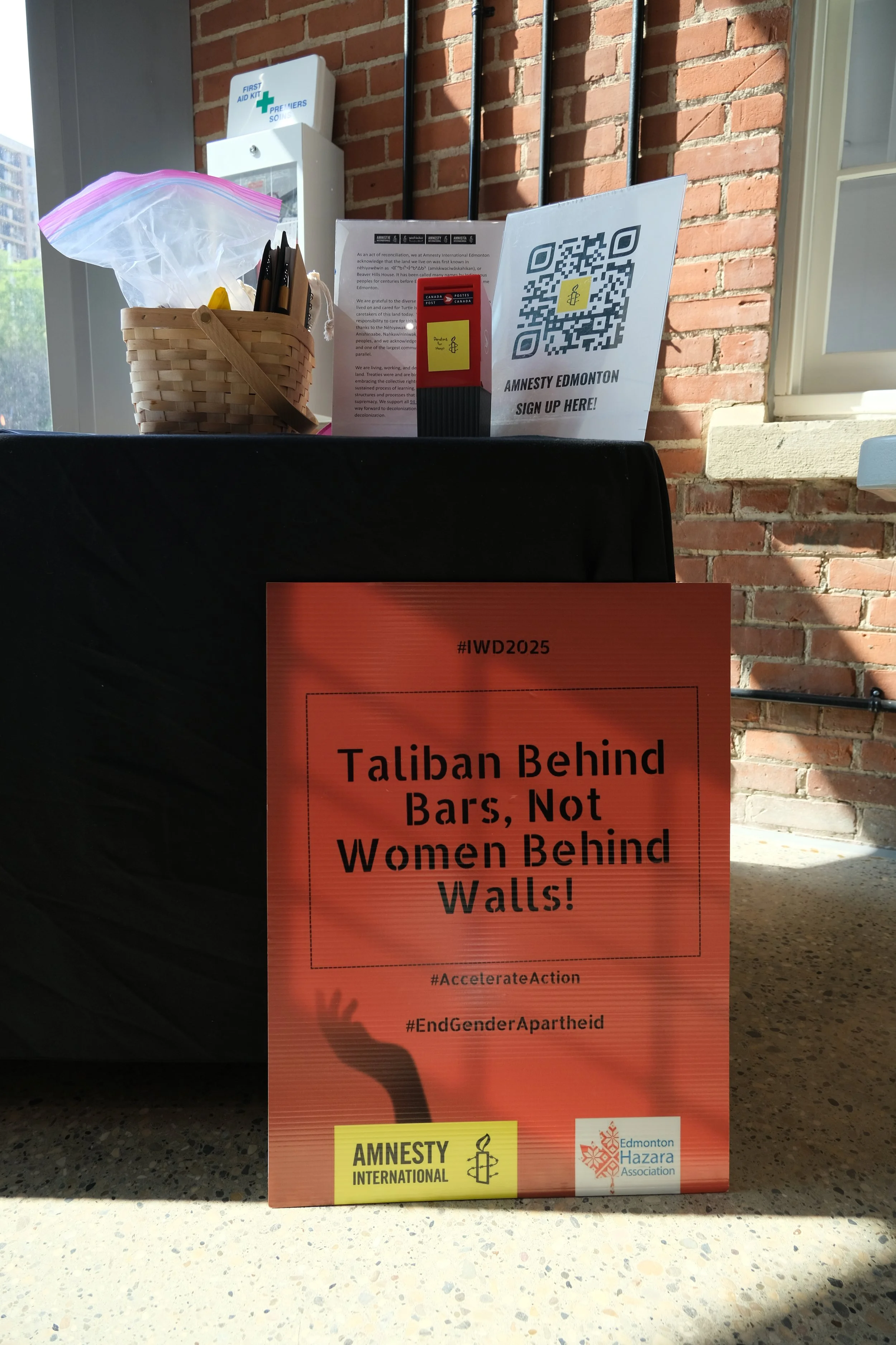 A red protest sign with the message 'Taliban Behind Bars, Not Women Behind Walls!' and hashtags #AccelerateAction and #EndGenderApartheid, along with logos of Amnesty International and Edmonton Hazara Association, placed on a countertop.