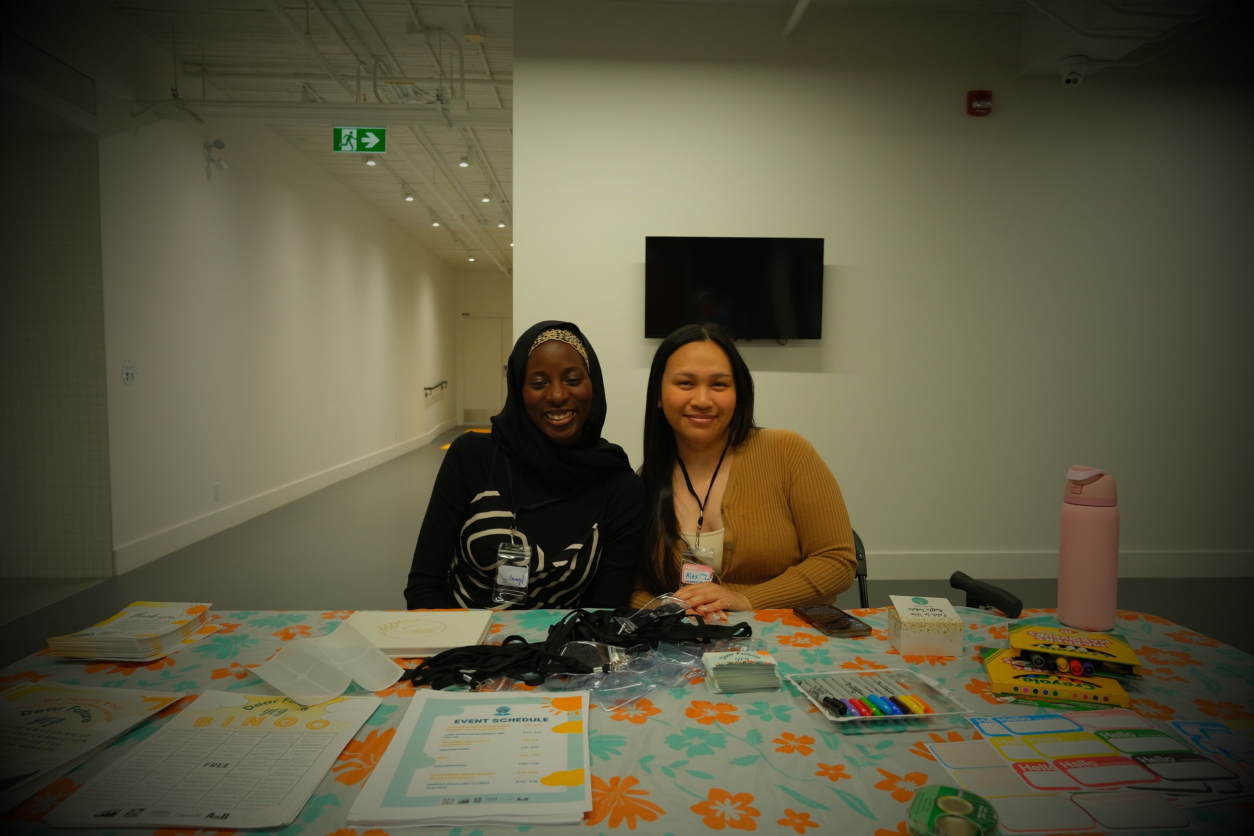 Two women sitting at a table with event materials, smiling for the camera in an indoor setting.