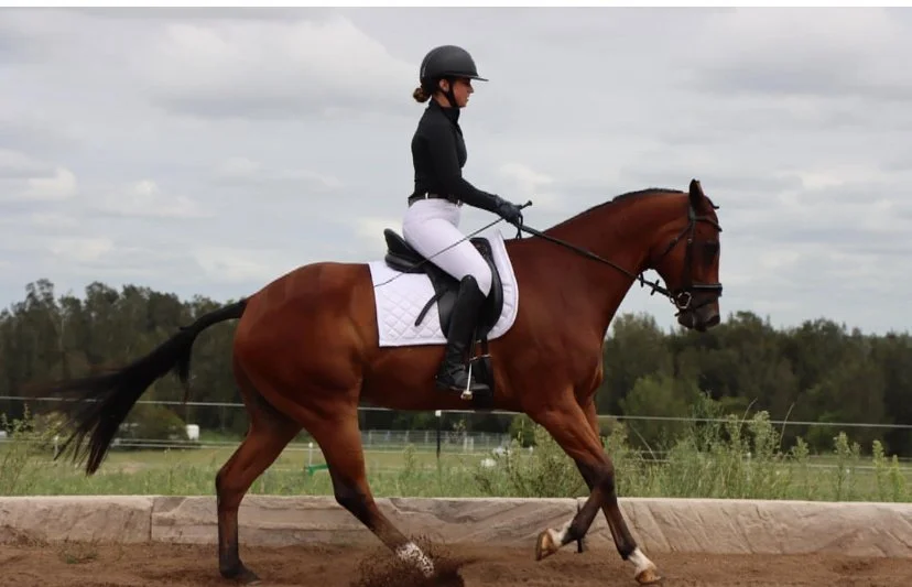 A woman riding a horse on a dirt track, wearing a black helmet, black jacket, white riding pants, and black riding boots, with green trees and cloudy sky in the background.