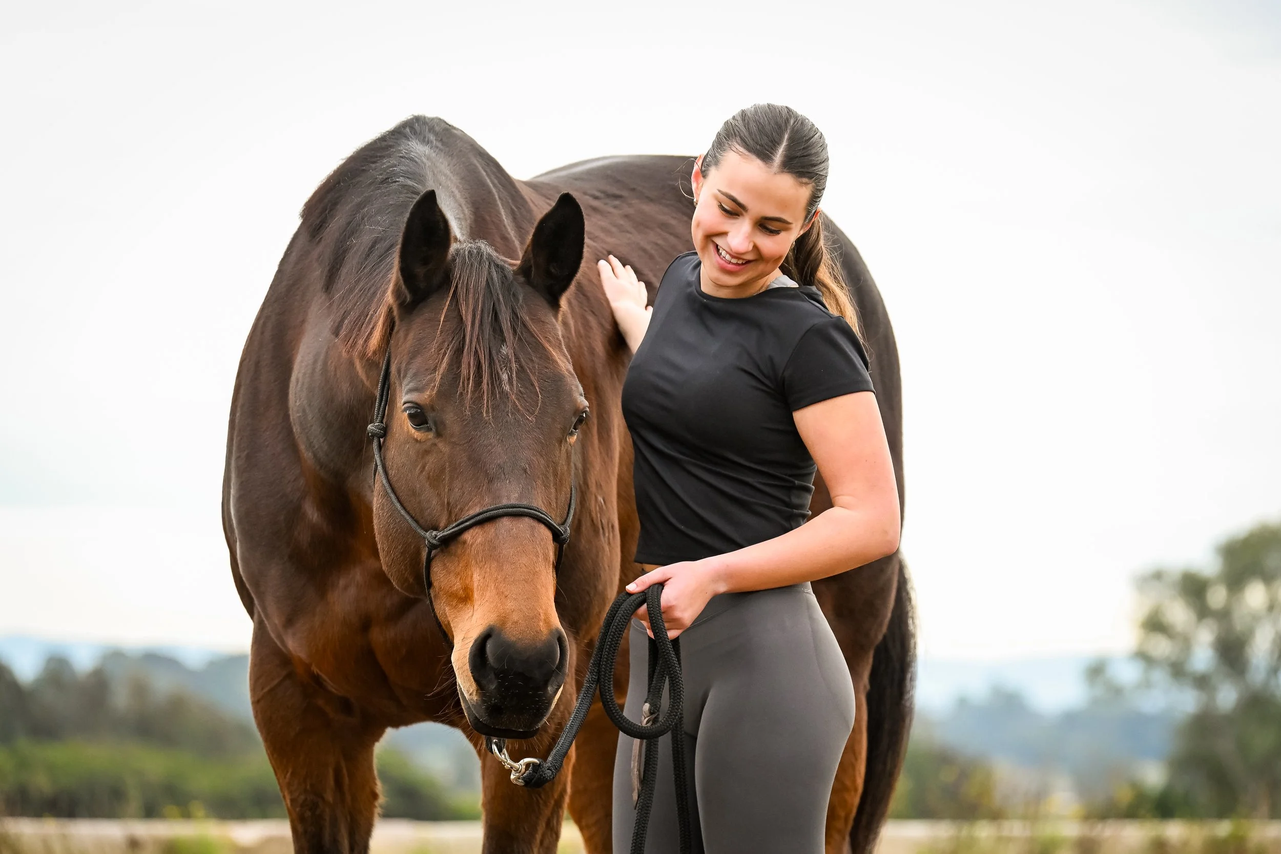 A woman in a black shirt and gray pants holding a lead rope stands next to a brown horse on a grassy field with trees and a cloudy sky in the background.
