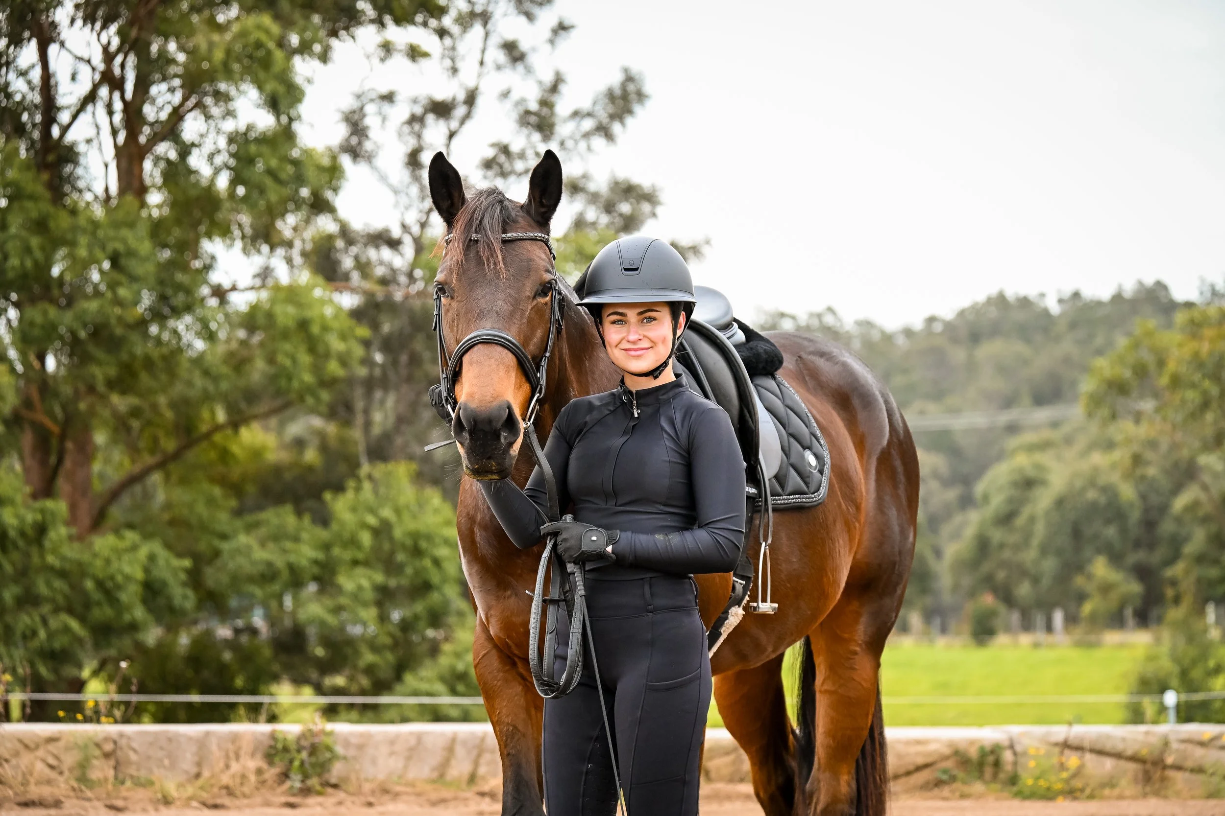 A woman in riding gear stands next to her brown horse with a bridle and saddle, outdoors in a green, wooded area.