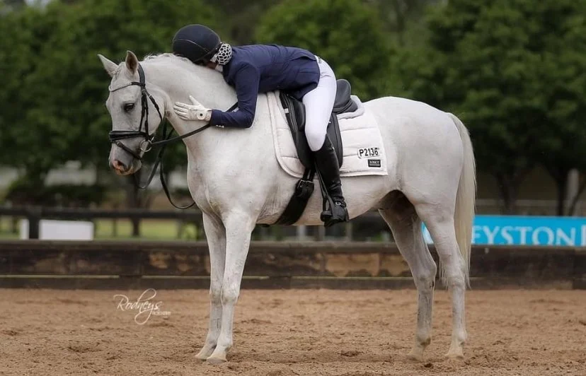 A jockey wearing a dark blue jacket, white riding pants, and a black helmet leans over and kisses a white horse on the neck while sitting on a saddle in an outdoor riding arena.