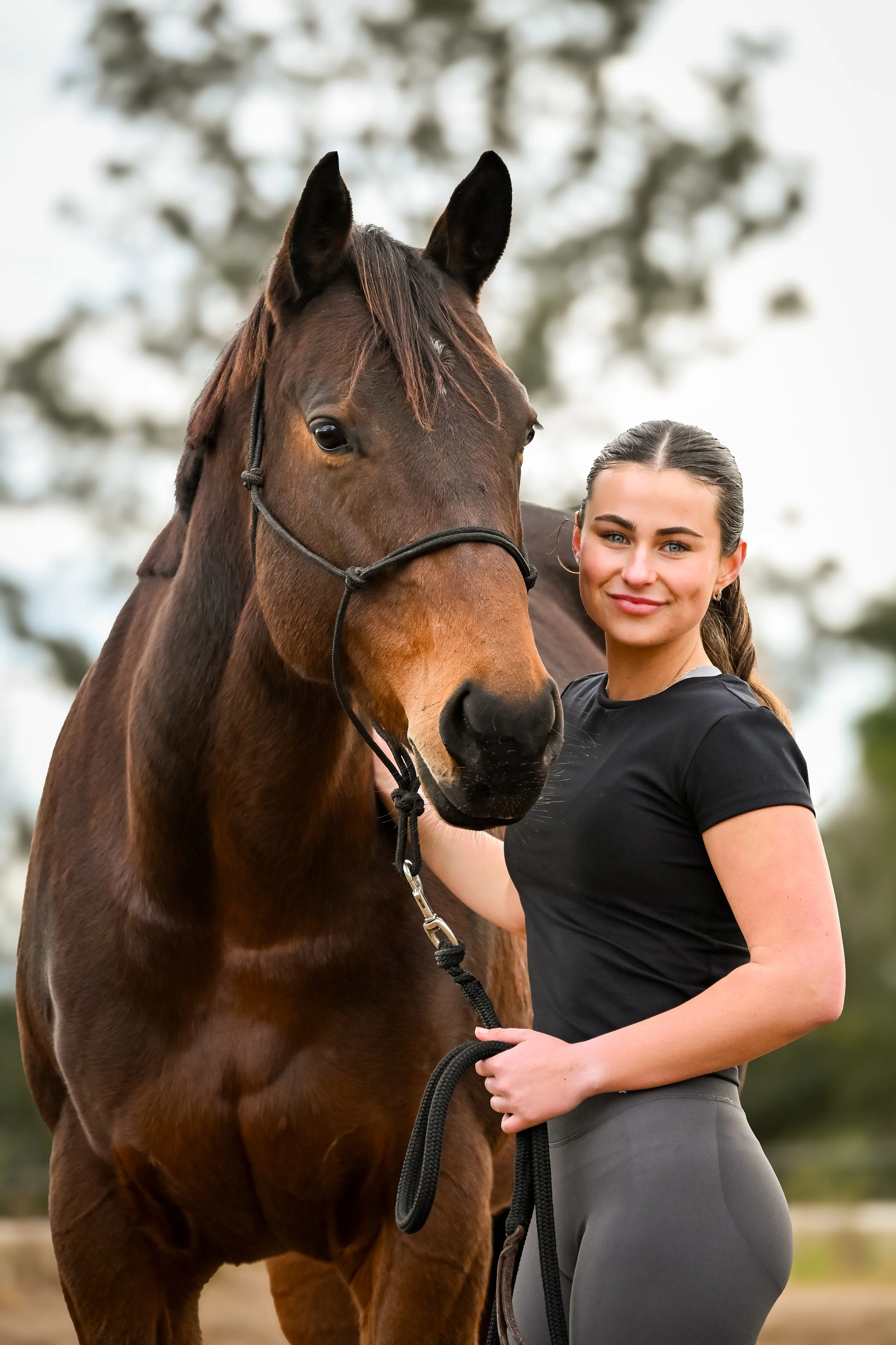A woman standing next to a brown horse outdoors, smiling, with trees in the background.
