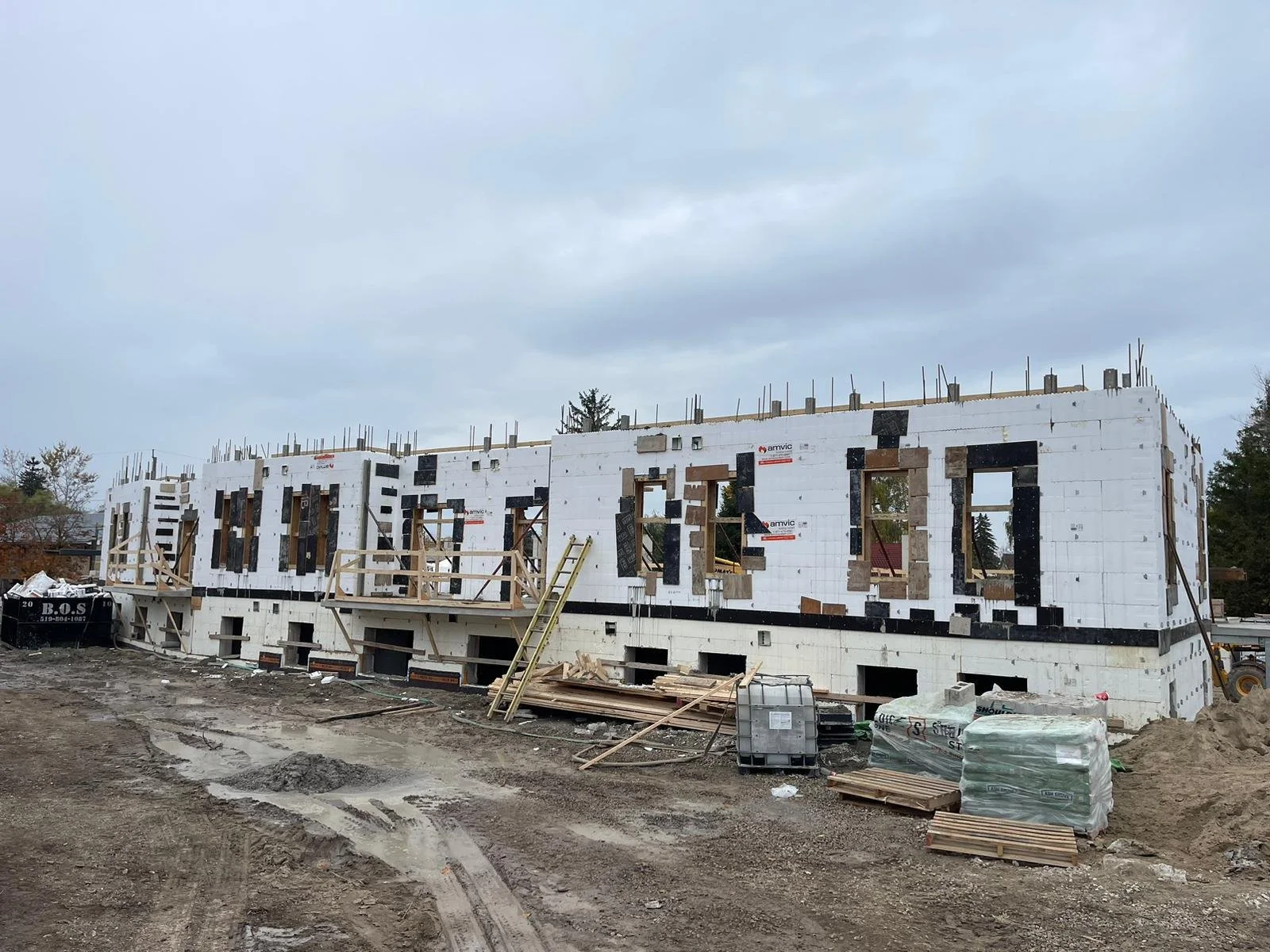 Construction site with a partially built multi-story building made of concrete blocks, with window openings and construction materials scattered around, under a cloudy sky.