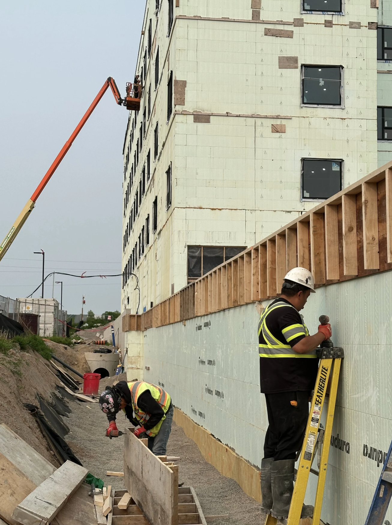 Construction workers building a multistory building. One worker is working on framing near the ground, while others are using a lift to work on the upper floors. The building's exterior wall is partially insulated and has windows installed.