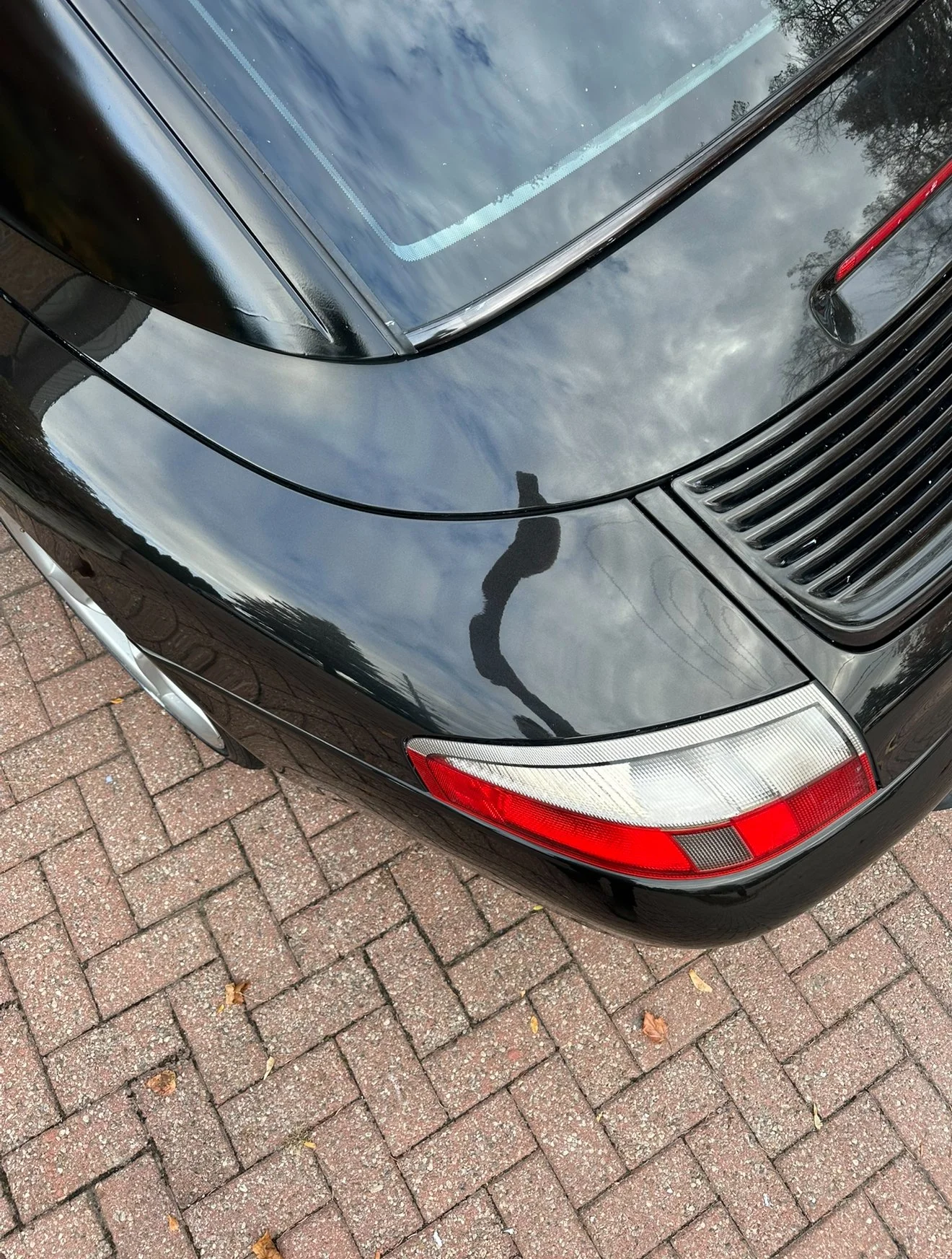 Close-up of the front right corner of a black car, showing the headlight, part of the windshield, and reflection of a person holding a phone, with a brick pavement underneath.