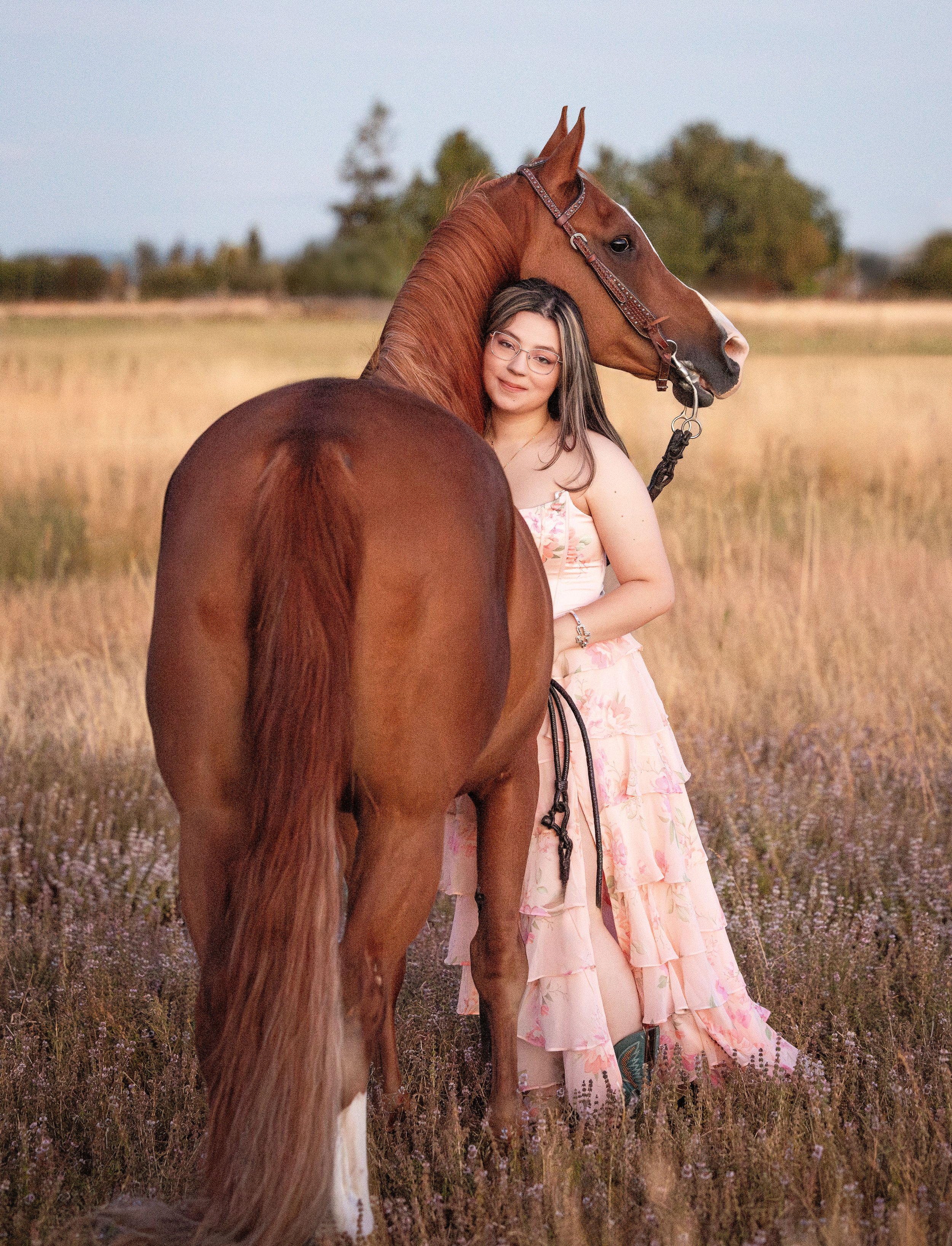 A woman with glasses and a floral dress standing in a field with a brown horse. The woman is smiling and looking at the horse, and the horse's head is resting against her head. The scene is set during the daytime with a blurred background of trees and grass.