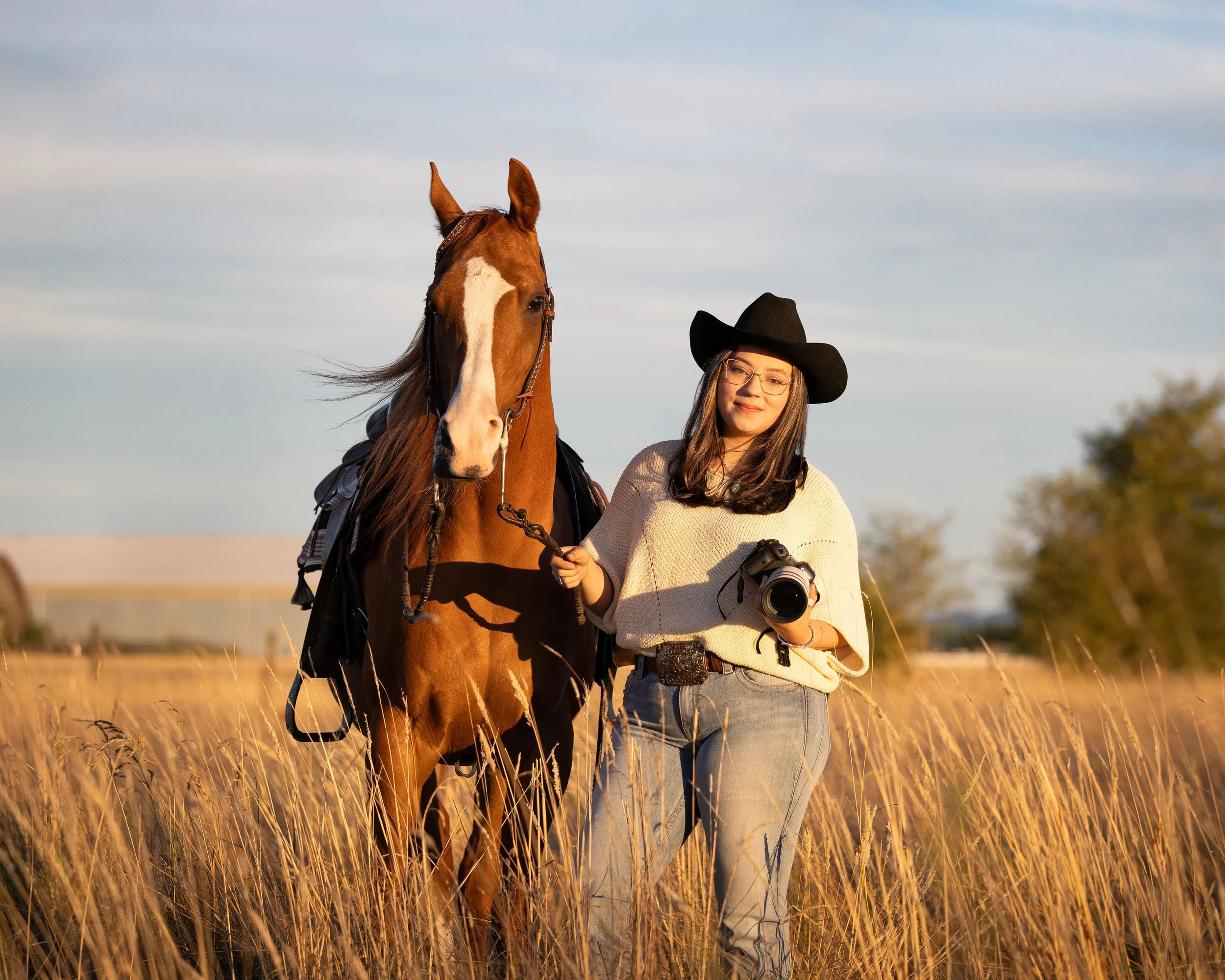 A woman wearing glasses and a black cowboy hat stands in a field of tall, golden grass with a horse. She is holding the horse's reins and has a camera hanging from her neck. The horse has a chestnut coat with a white stripe down its face and is wearing a saddle and bridle. The sky is partly cloudy with a soft, warm light, suggesting late afternoon or early evening.