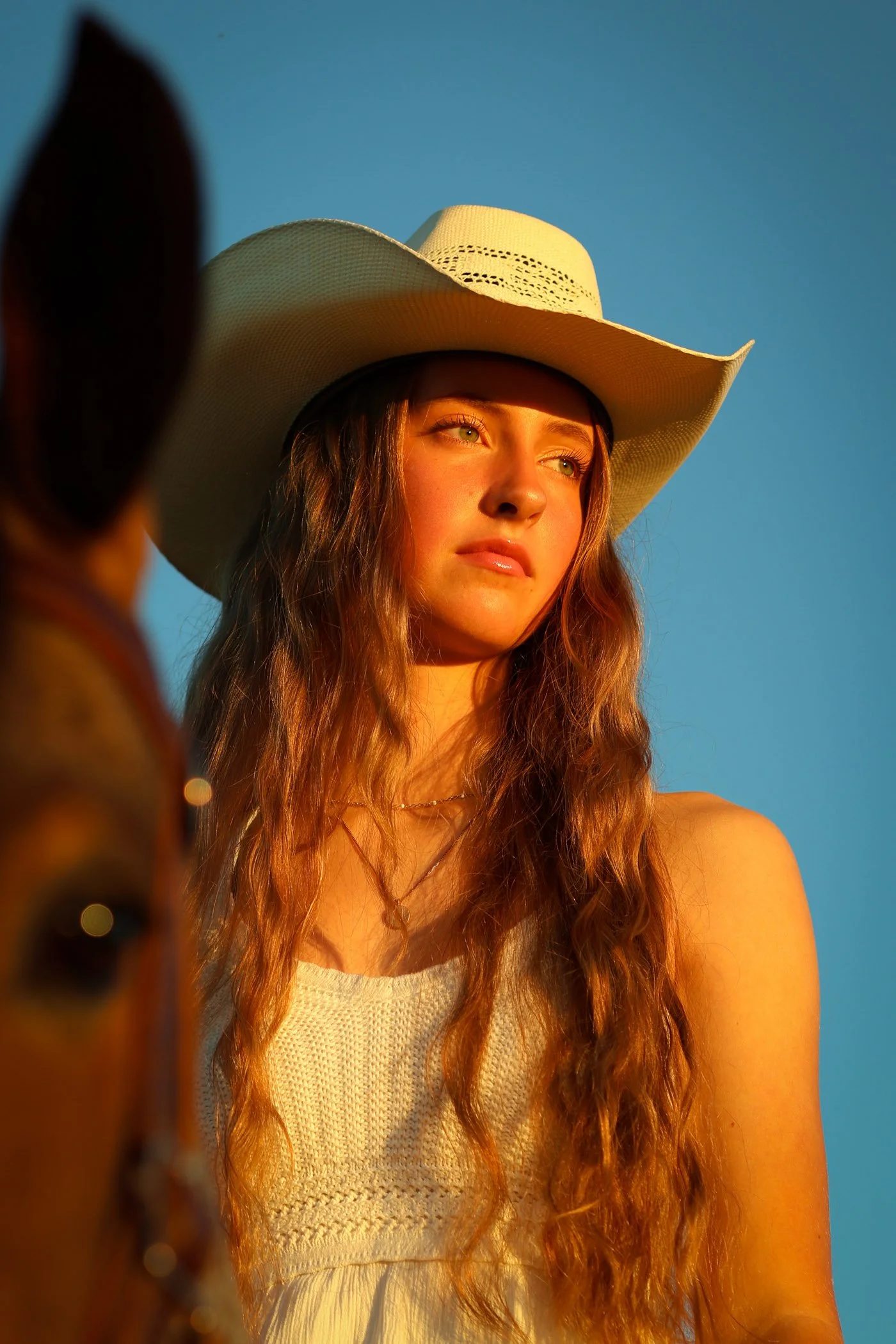 A young woman with long wavy hair wearing a white cowboy hat and sleeveless top, standing outdoors with a blue sky background.