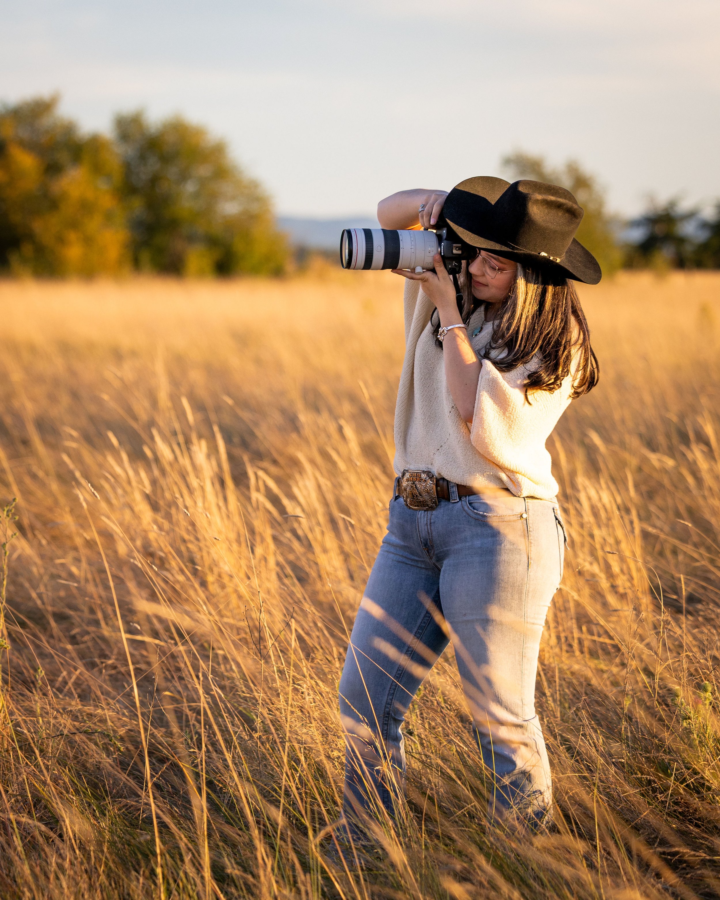 A woman wearing glasses, a large black hat, cream sweater, and jeans is standing in a field of tall golden grass, taking a photograph with a camera during sunset.