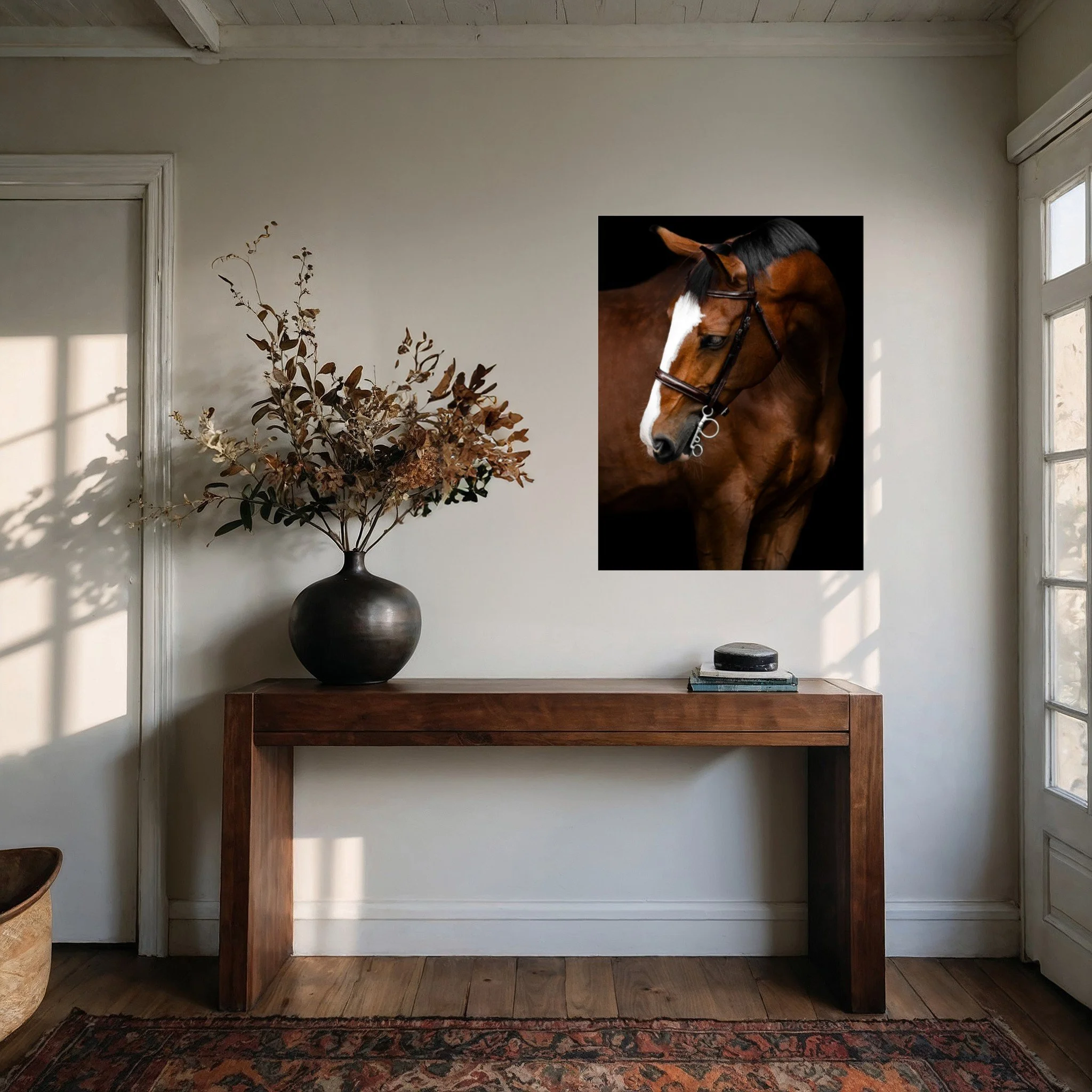 A room with a wooden console table, a black vase with dried leaves, a stack of books, and a large poster of a brown horse with a white stripe on its face hanging on the wall near windows.