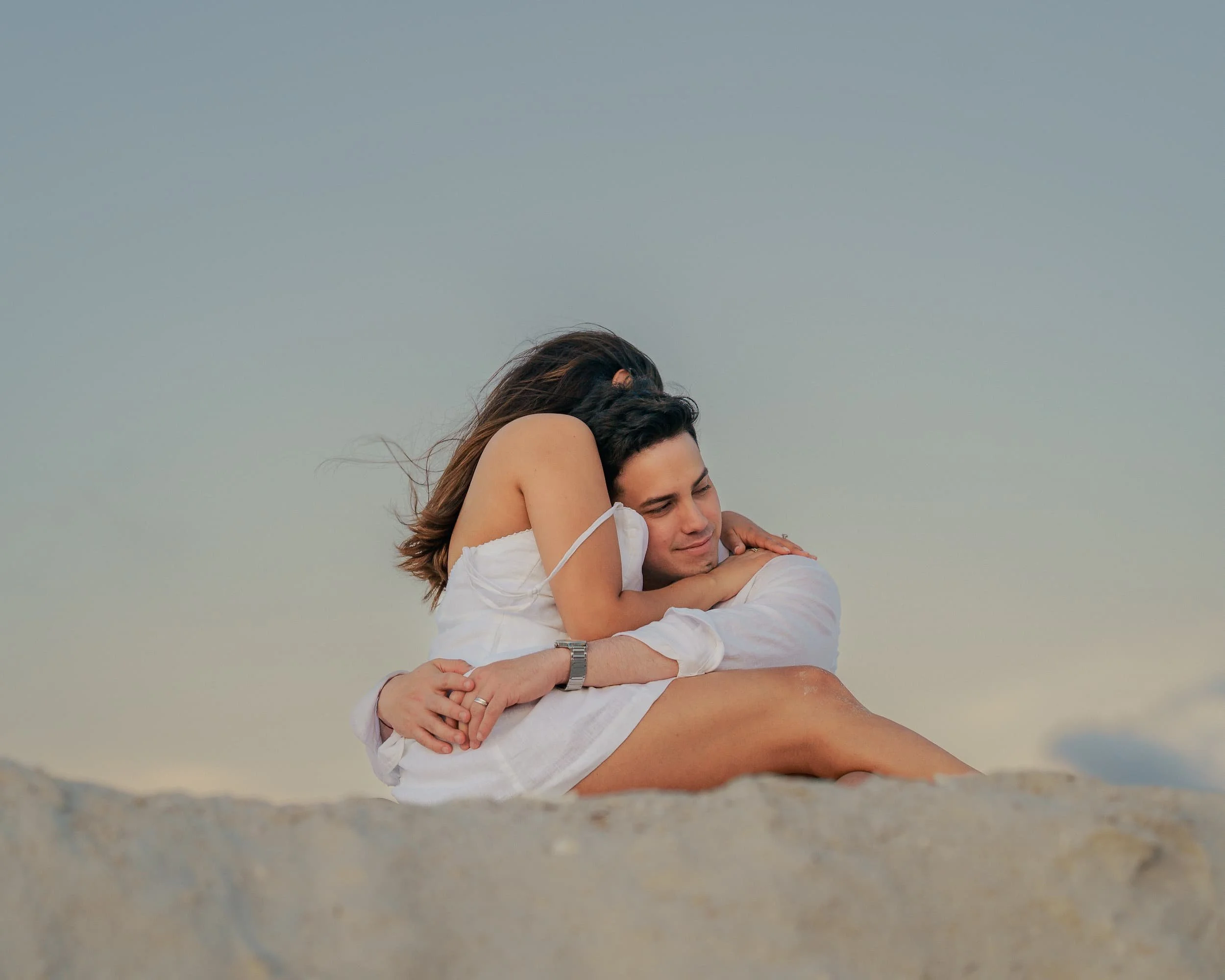 A couple sitting on a sandy surface, hugging and smiling with a clear sky background. Cinematic Editorial Pictures in Tampa Florida.  Engagement pictures in florida, tampa.