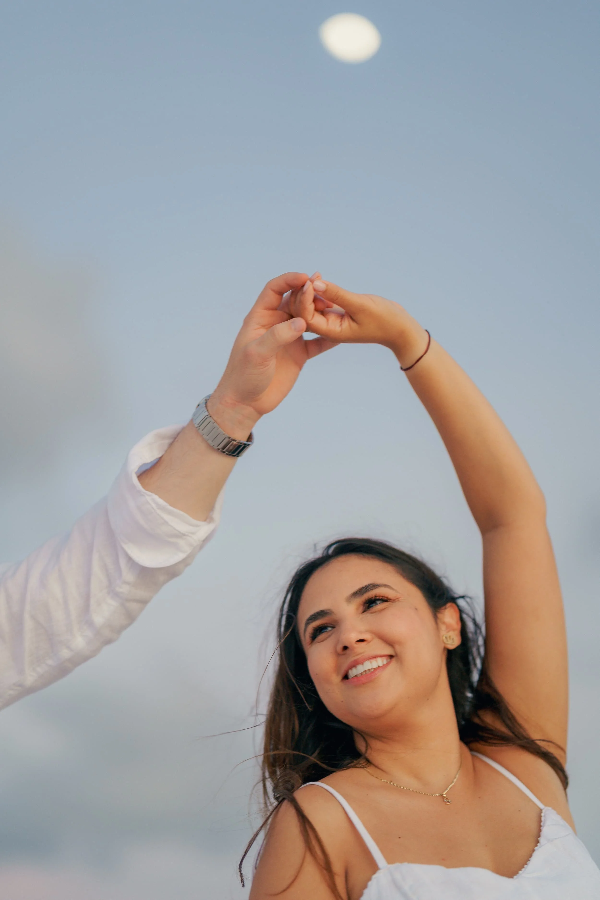 A young woman with long brown hair wearing a white sleeveless top, smiling and looking at the sky, as a person with a watch and a white shirt holds her hand and forms a heart shape against a background of a cloudy sky with the moon visible. Cinematic
