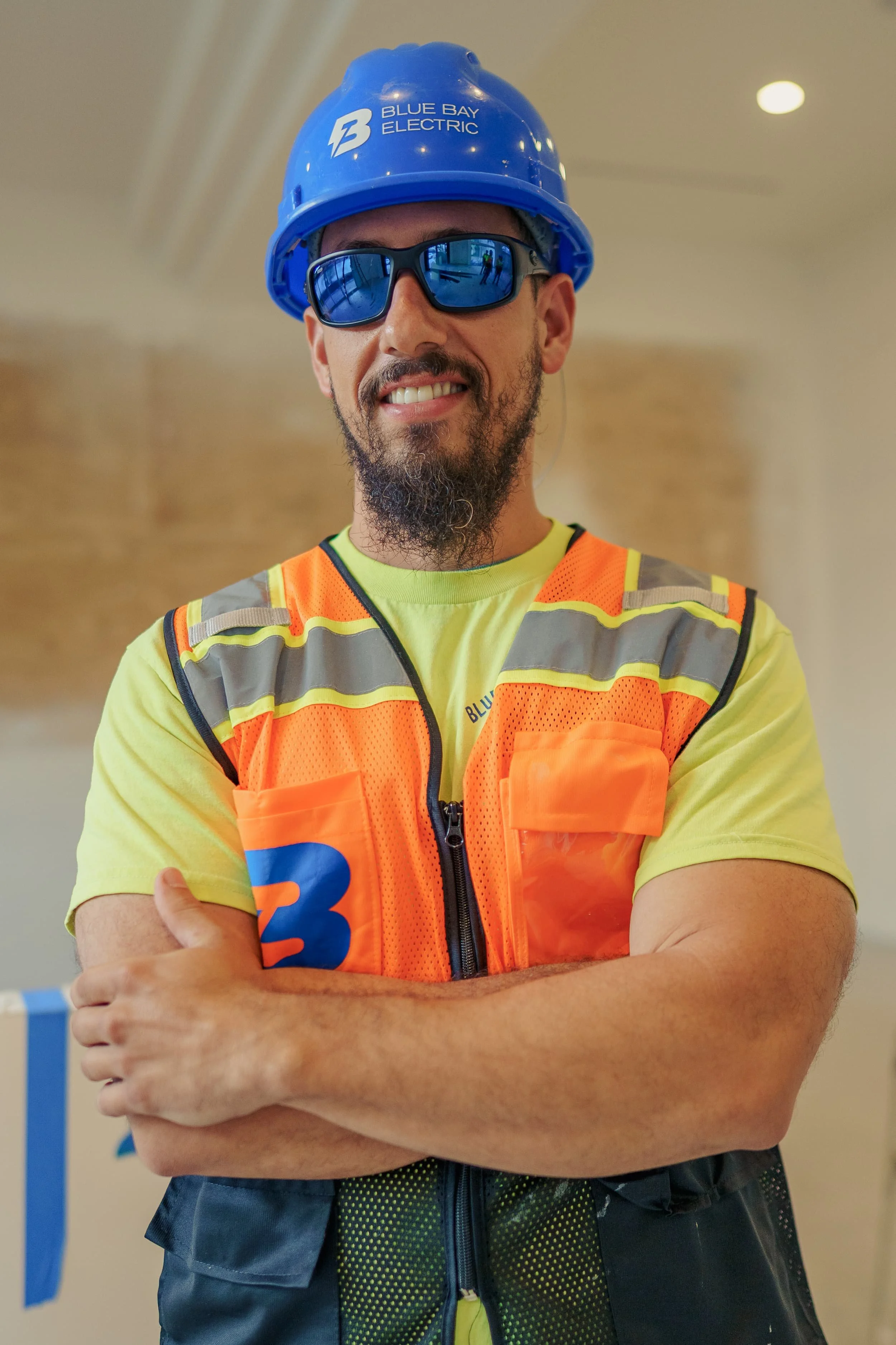 A construction worker wearing a blue hard hat with "Blue Bay Electric" logo, reflective sunglasses, a neon yellow T-shirt, and an orange safety vest with reflective stripes, standing indoors with arms crossed and smiling. Cinematic Editorial Pictures