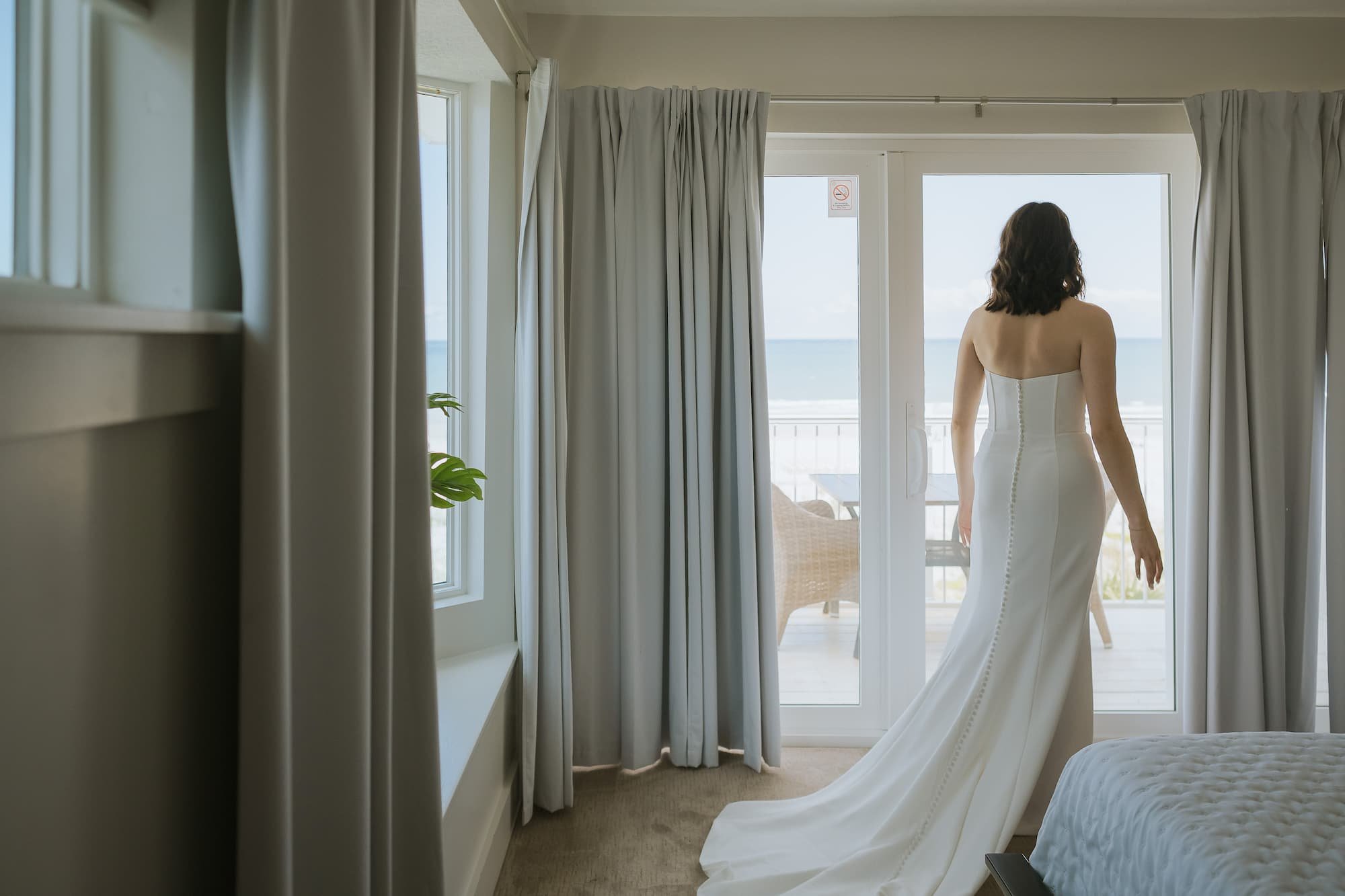 A woman in a white strapless wedding gown standing on a balcony, looking out at the ocean.
